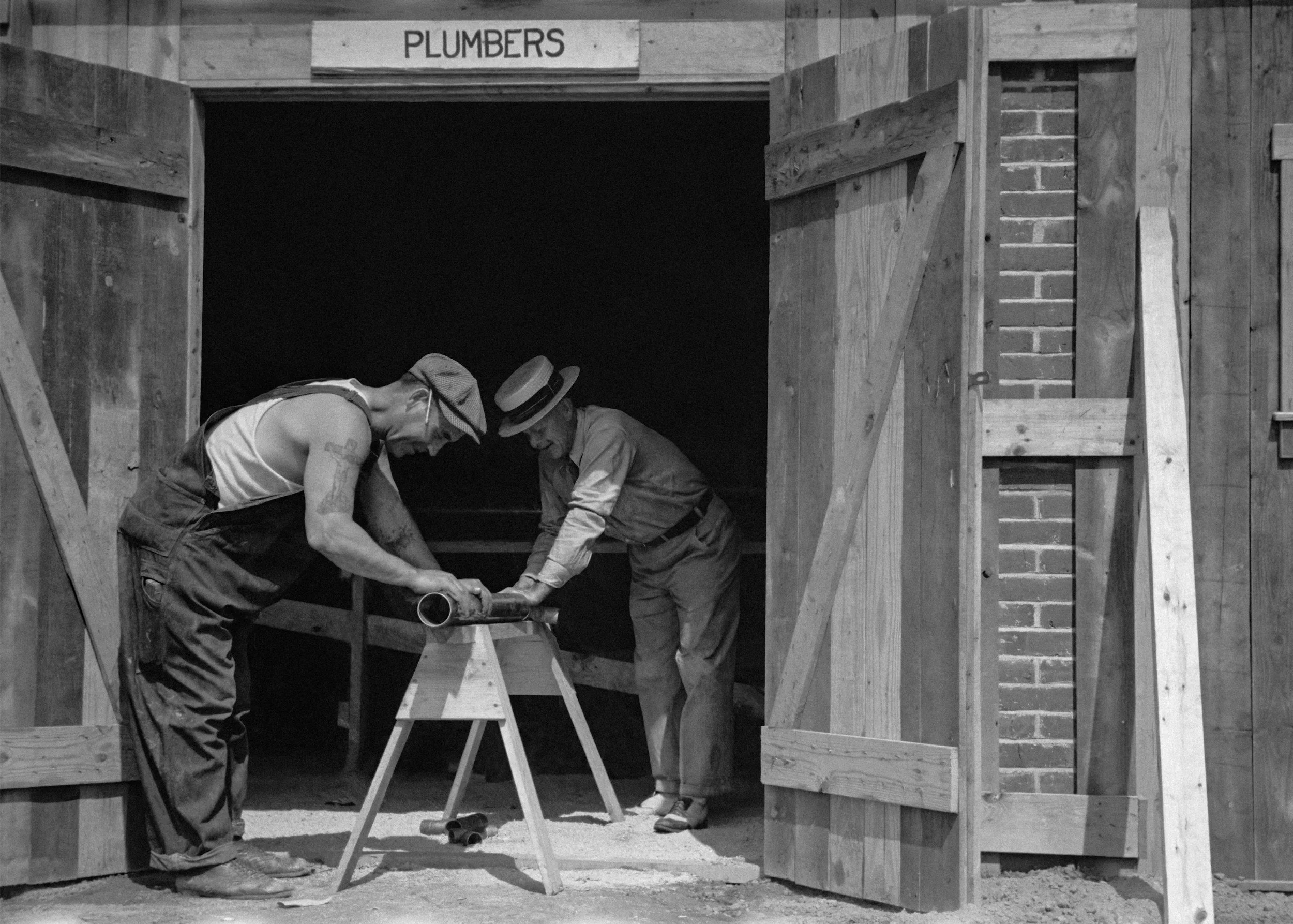 Two construction workers, both wearing hats and safety glasses, working together on a wooden structure labeled "PLUMBERS" in a workshop or construction site.