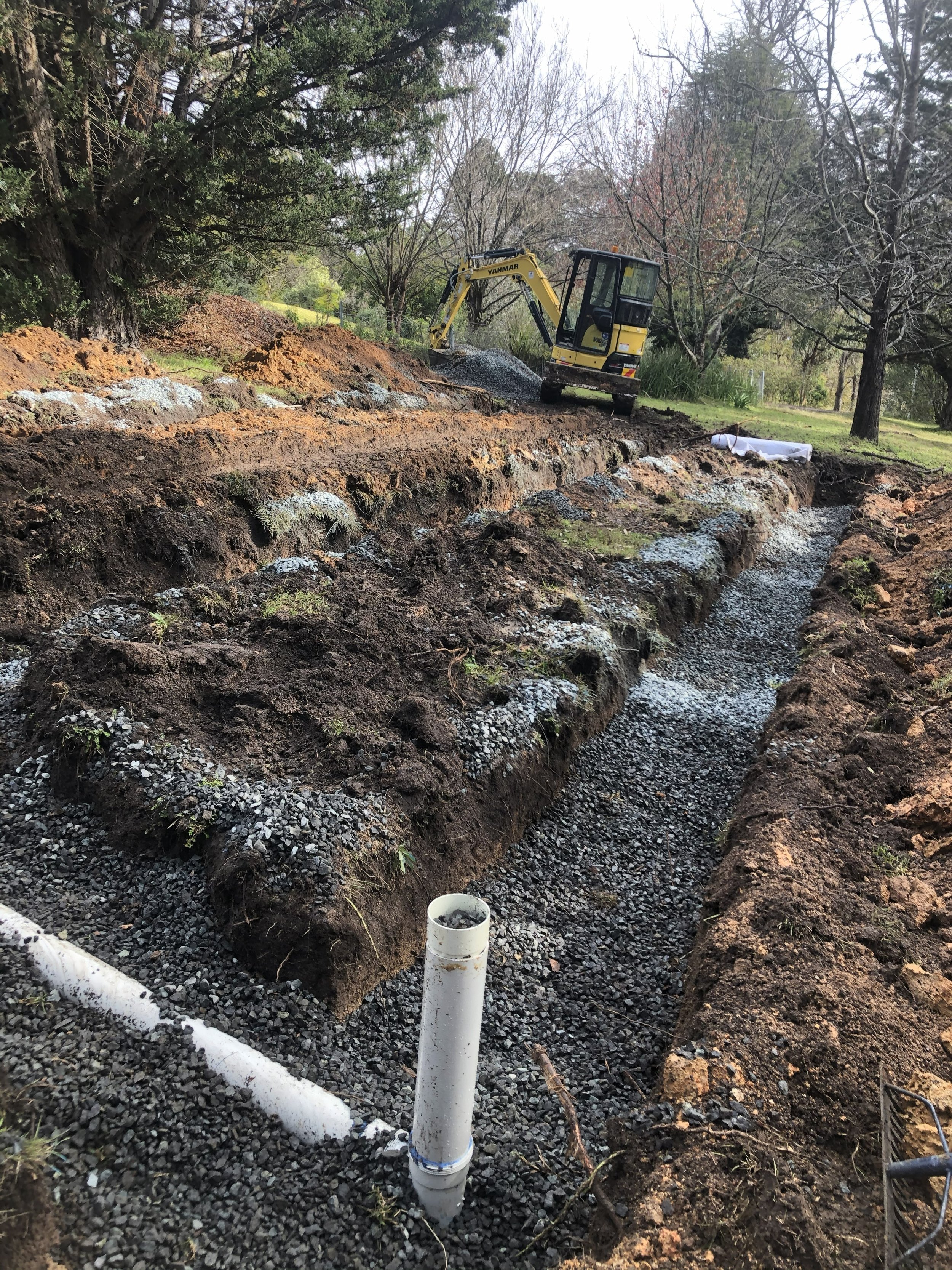 A construction site in a backyard with a small excavator digging a trench. There are trees and grass surrounding the area, and white pipes are visible in the excavated trench.