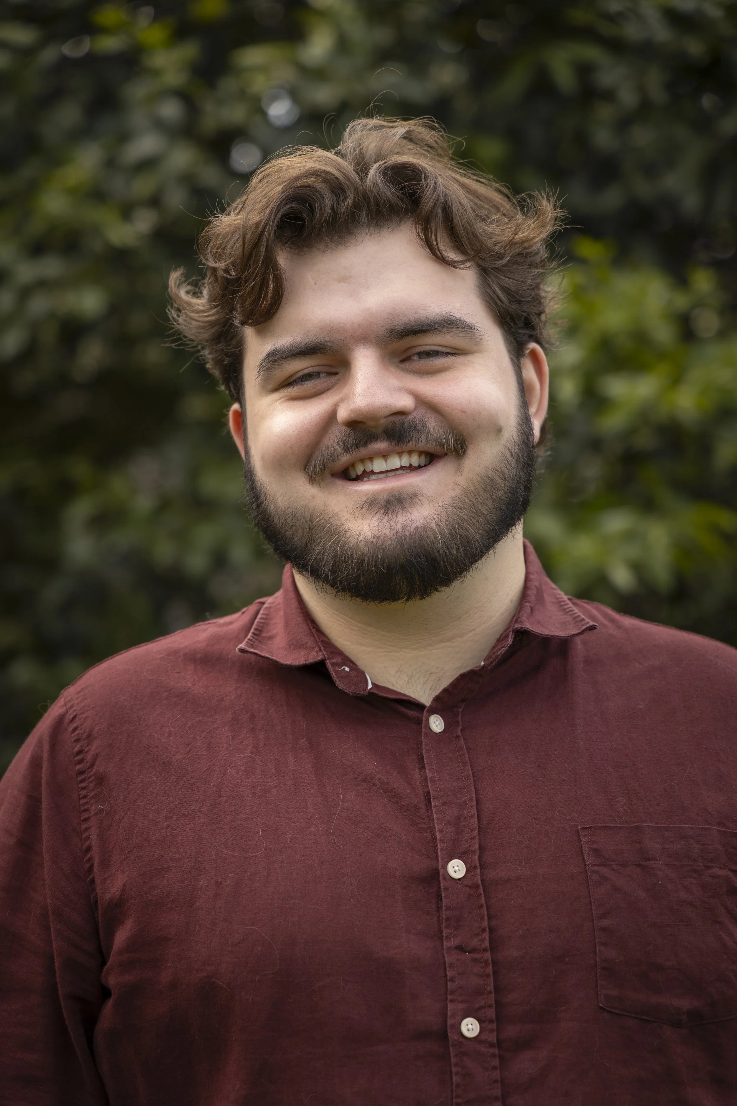 A young man with light skin, brown curly hair, and a beard smiling outdoors, wearing a maroon button-up shirt, with a background of blurred green foliage.
