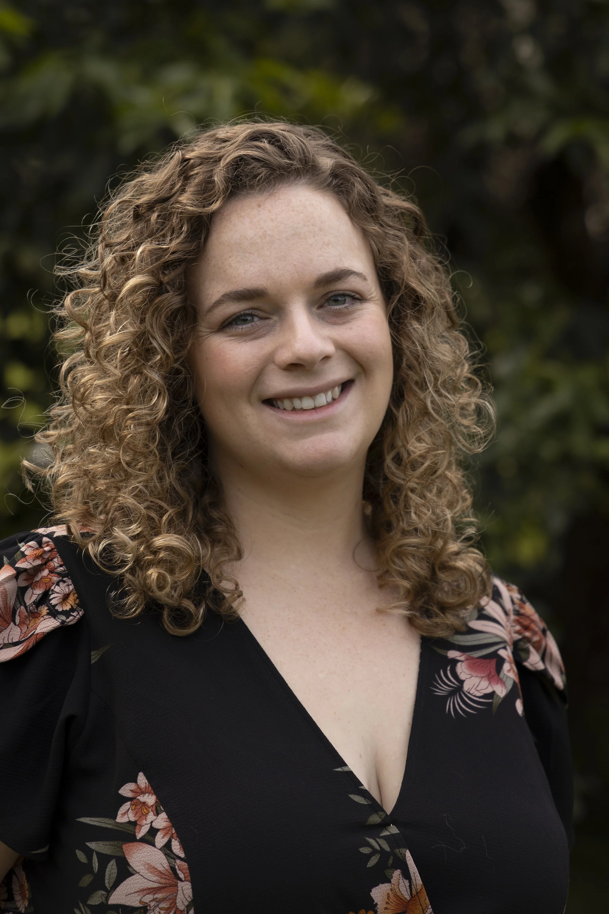 A smiling woman with curly blonde hair, wearing a black dress with floral patterns, standing outdoors with greenery in the background.