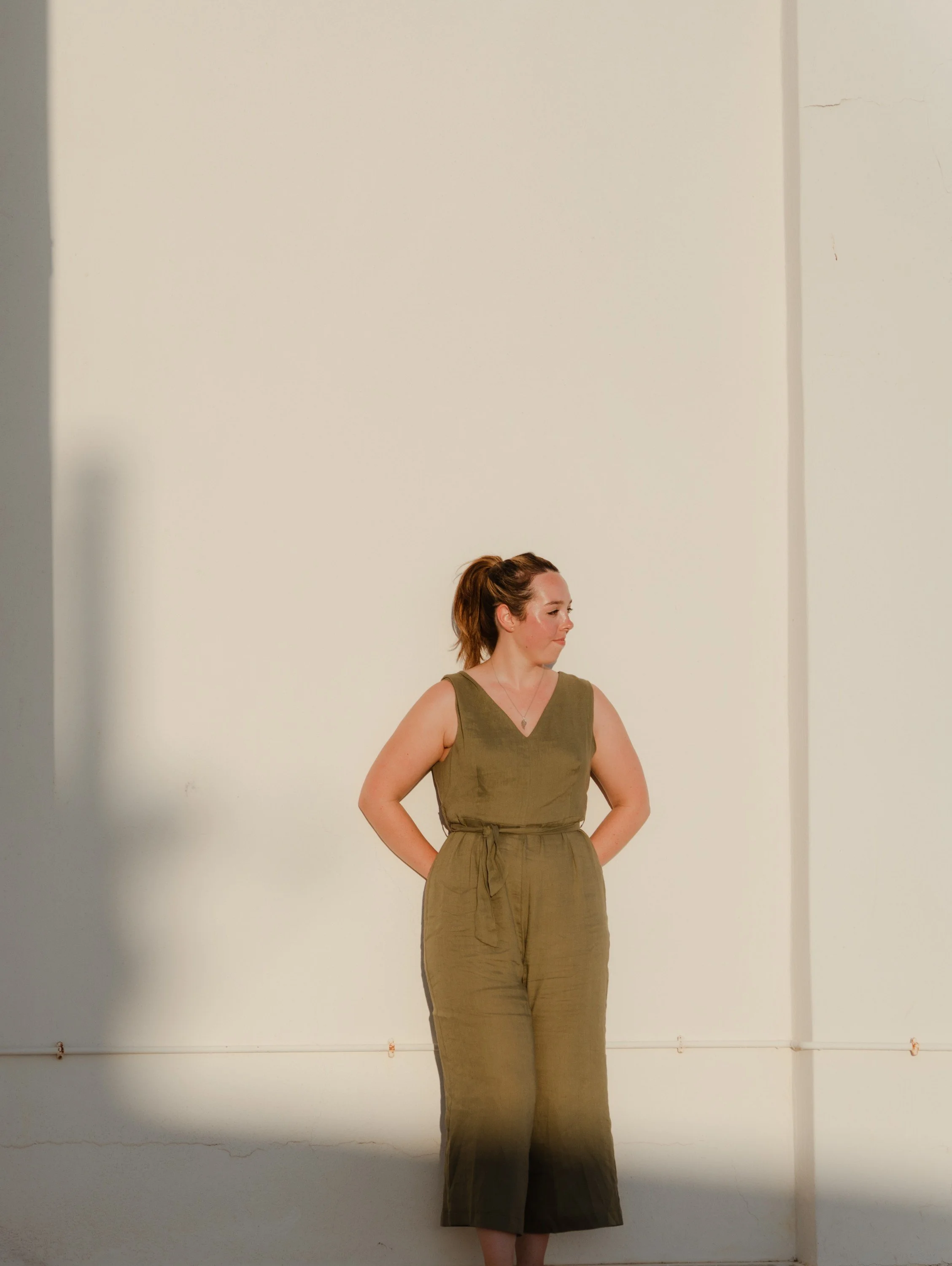 A woman with brown hair tied in a ponytail, wearing a sleeveless olive green jumpsuit, standing against a plain white wall with a small crack and some rust marks at the bottom.