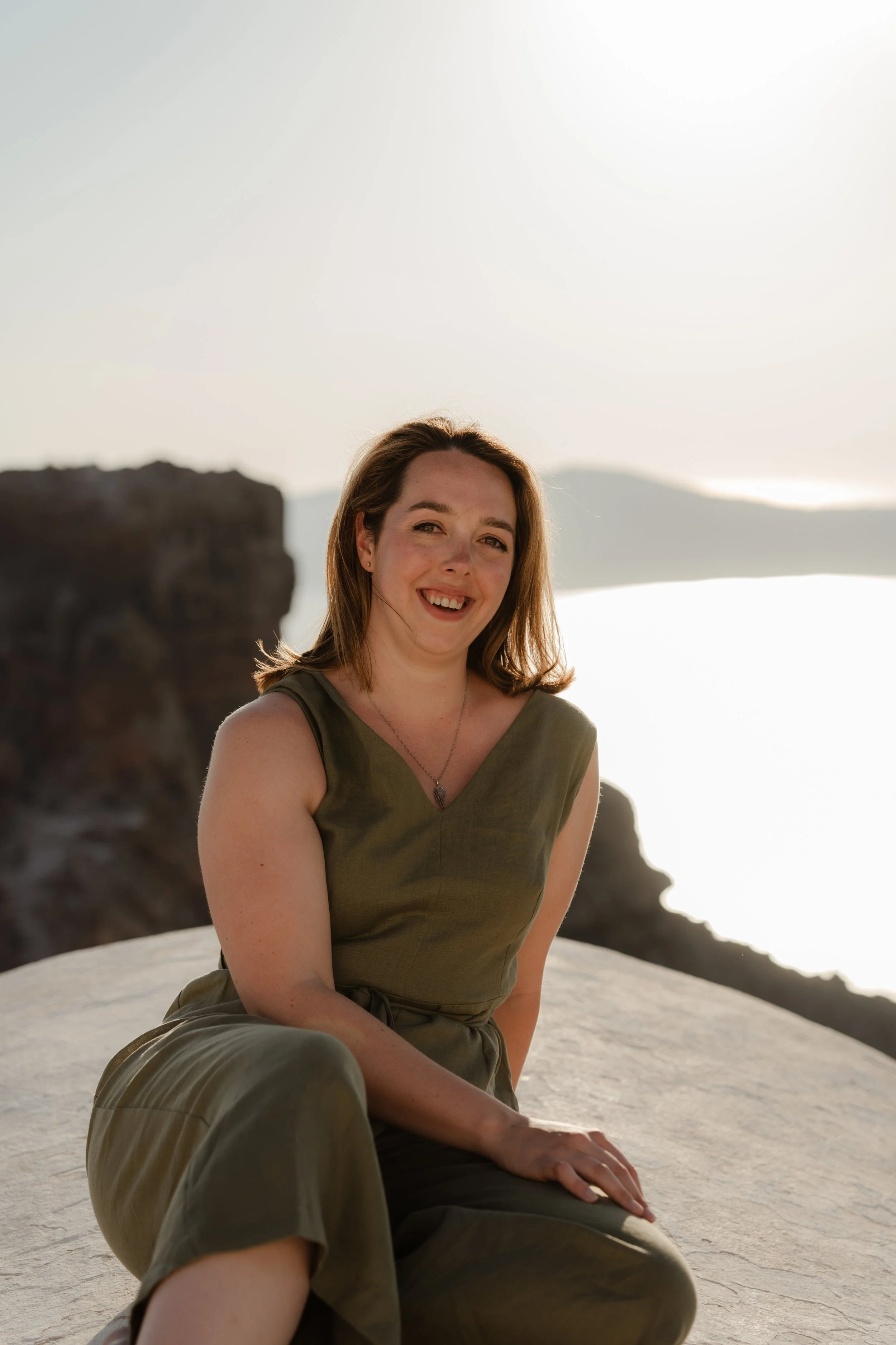 A woman with shoulder-length brown hair smiling, sitting on a large rock formation outdoors during daytime with a distant mountain and cloudy sky in the background.