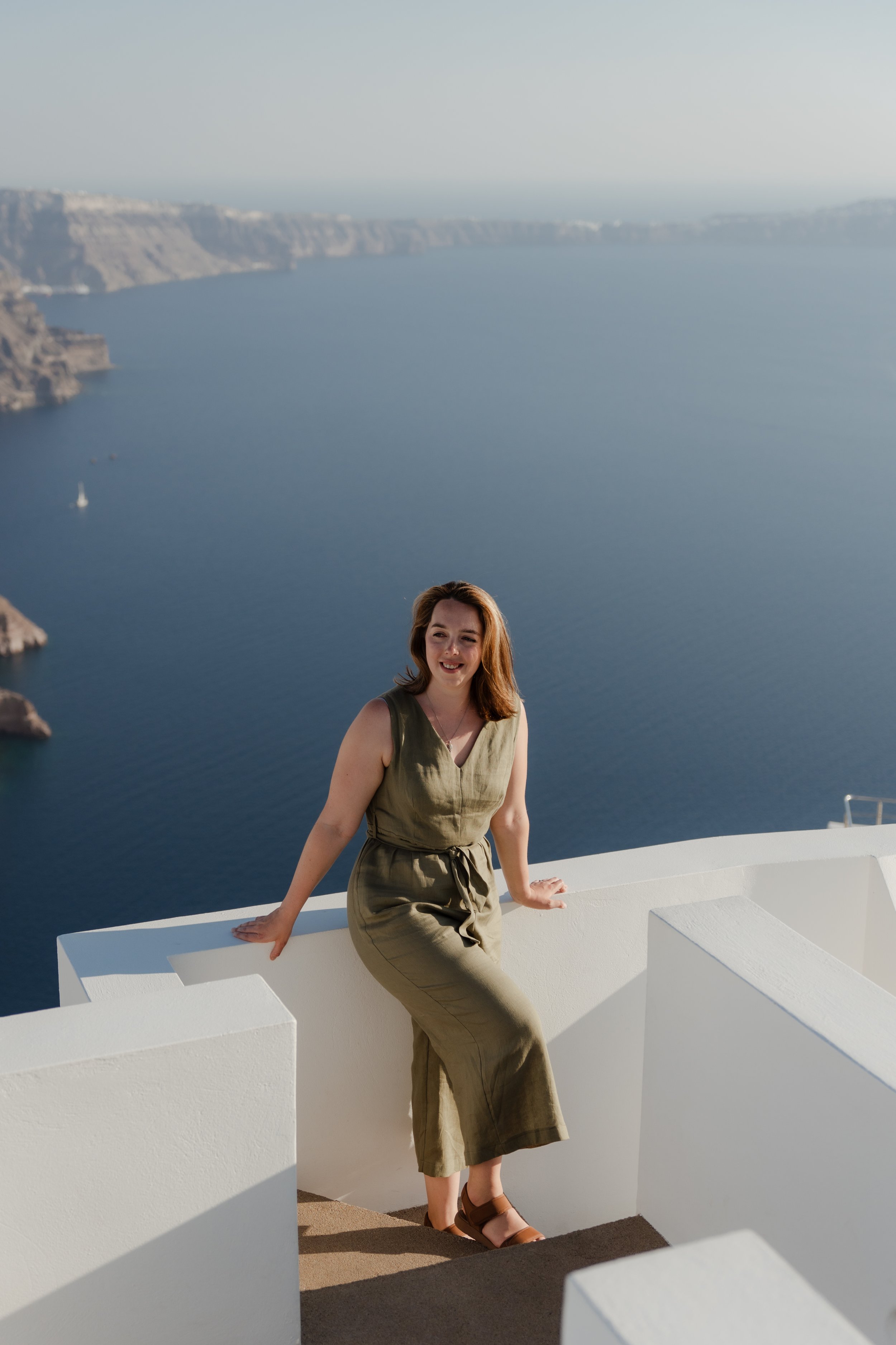 A woman with shoulder-length brown hair wearing a sleeveless khaki dress and sandals, standing and leaning on a white wall with a scenic view of the ocean, cliffs, and boats in the background.