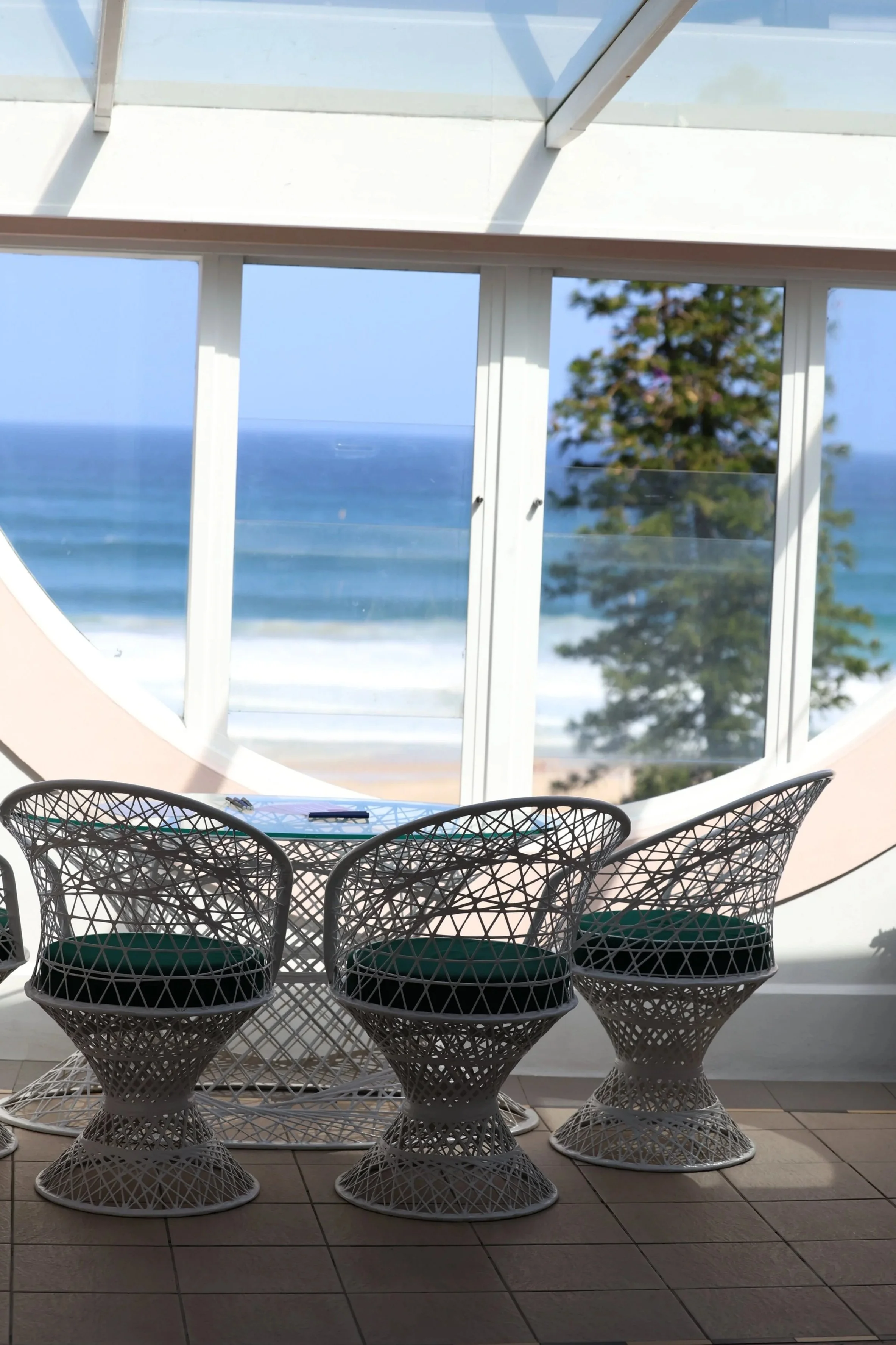 A set of white modern wire chairs with dark cushions inside a bright room with large windows showing the ocean, tree, and blue sky outside.