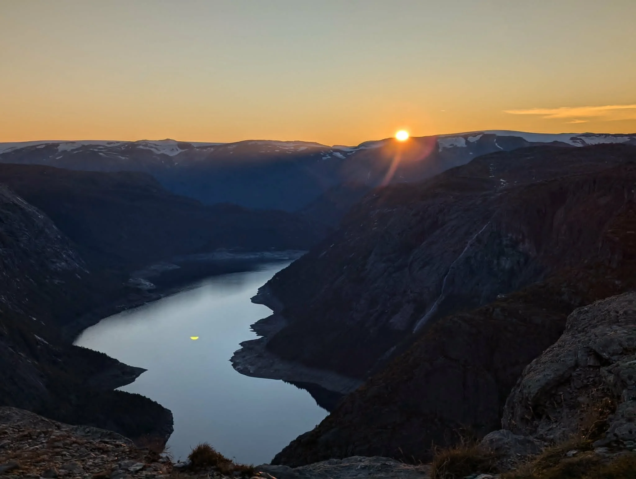 Sun setting behind snow-capped mountains over a deep valley with a winding river.