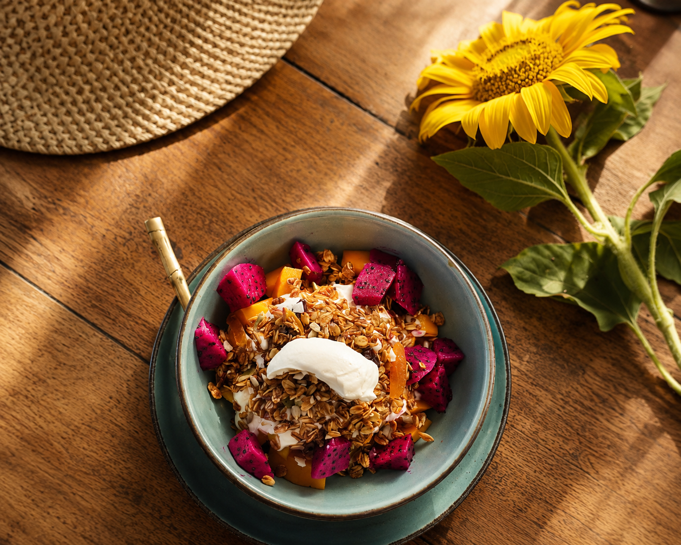 A bowl of yogurt topped with granola, sliced dragon fruit, and mango, with a spoon inside, on a wooden table next to a sunflower and a straw hat.