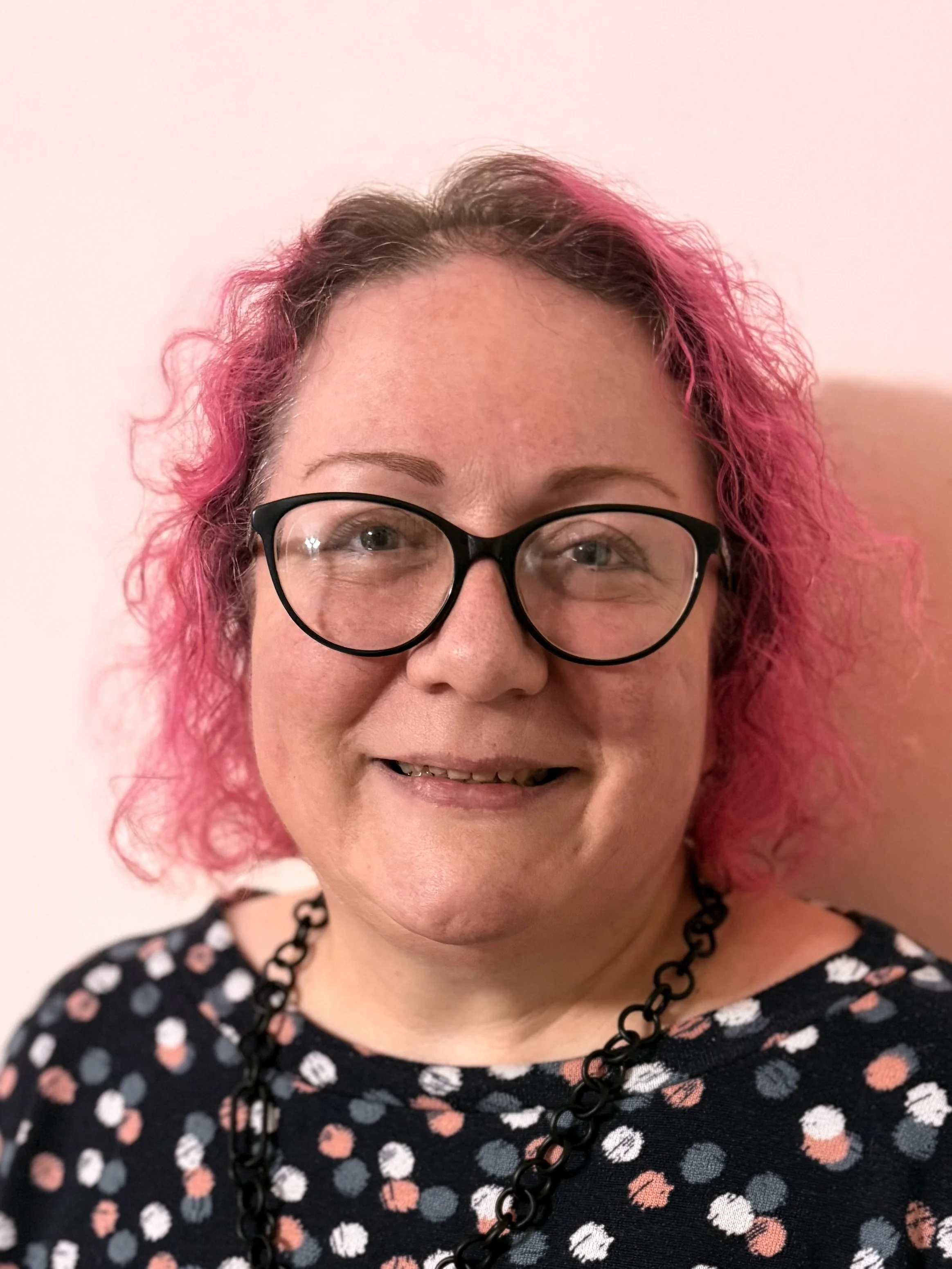A woman with curly pink and brown hair, wearing glasses and a black necklace, smiling in front of a light-colored wall.