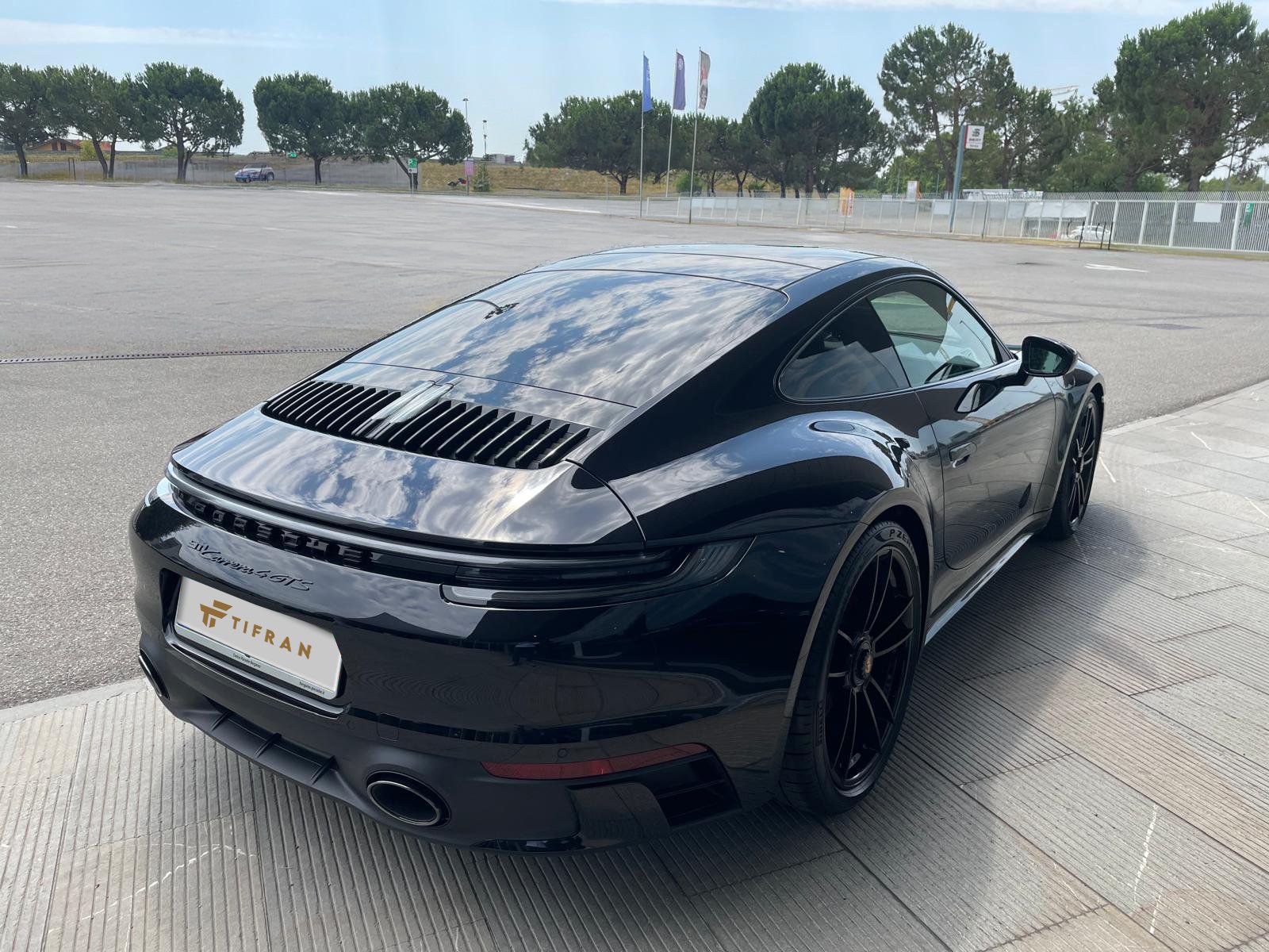 Porsche 911 Auto sportiva di colore nero parcheggiata su un marciapiede con alberi e bandiere sullo sfondo.