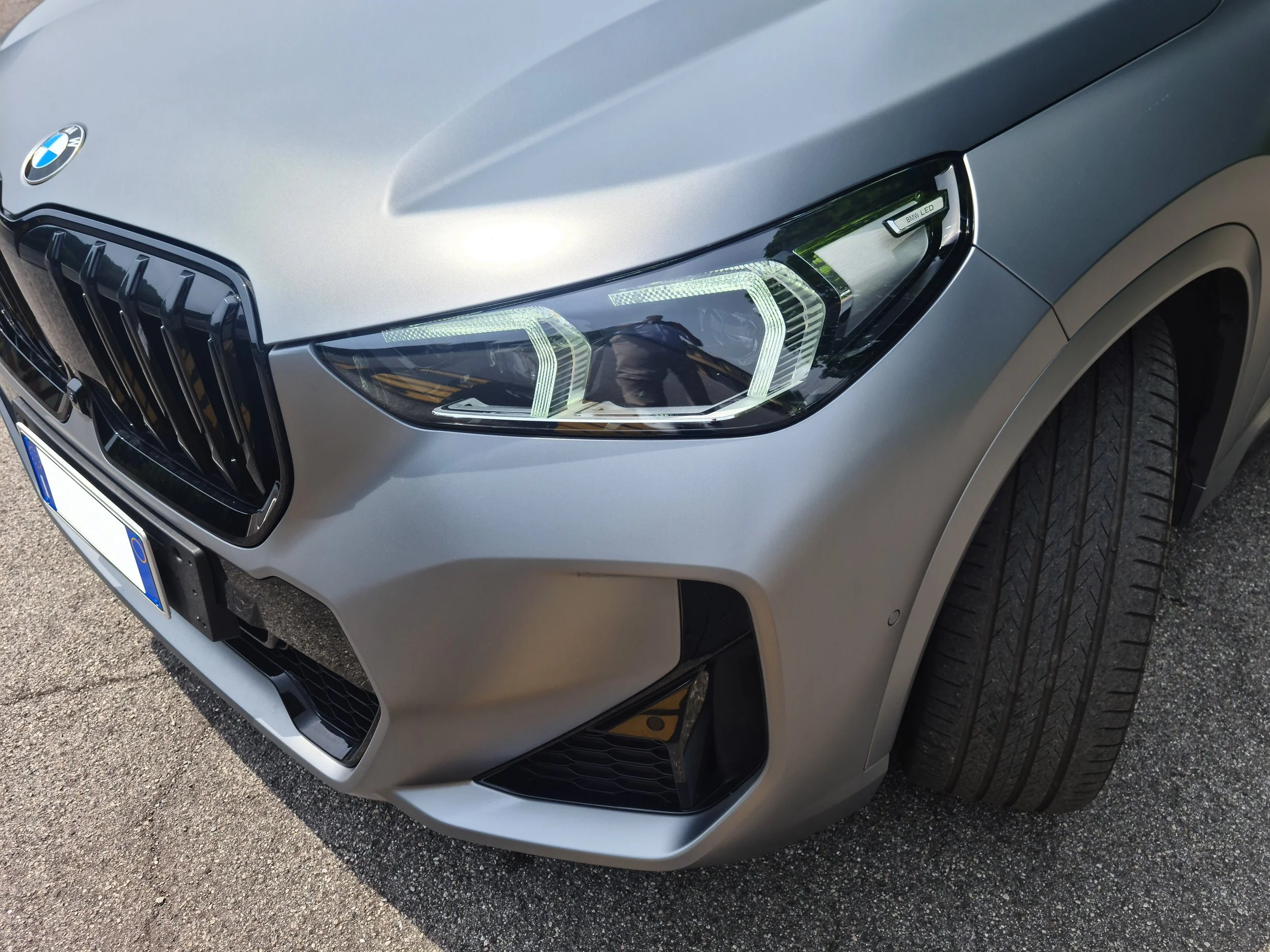 Prima parte di una macchina BMW grigia, vista dal davanti, con evidenti i fari anteriori a LED e la griglia caratteristica. La macchina sembra essere parcheggiata su una strada asfaltata.