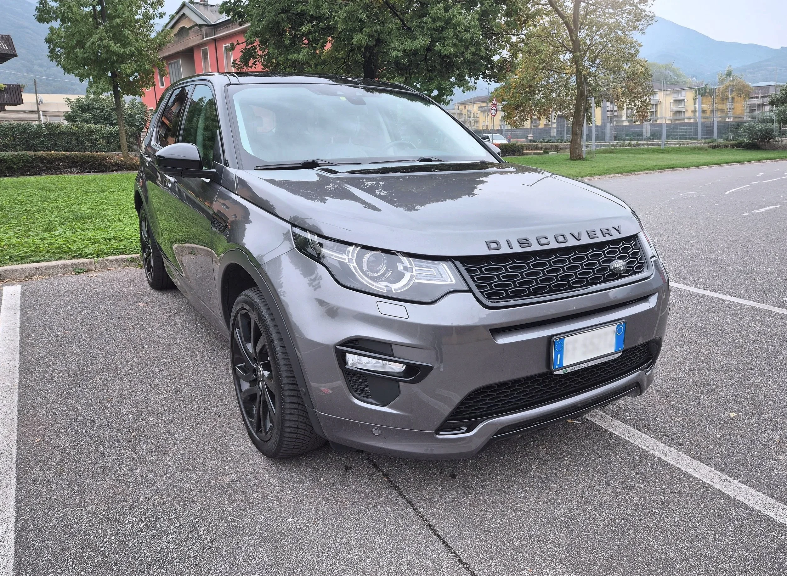 Auto SUV di colore grigio, marca Land Rover, modelloDiscovery, parcheggiata su una strada con sfondo di cespugli verdi.