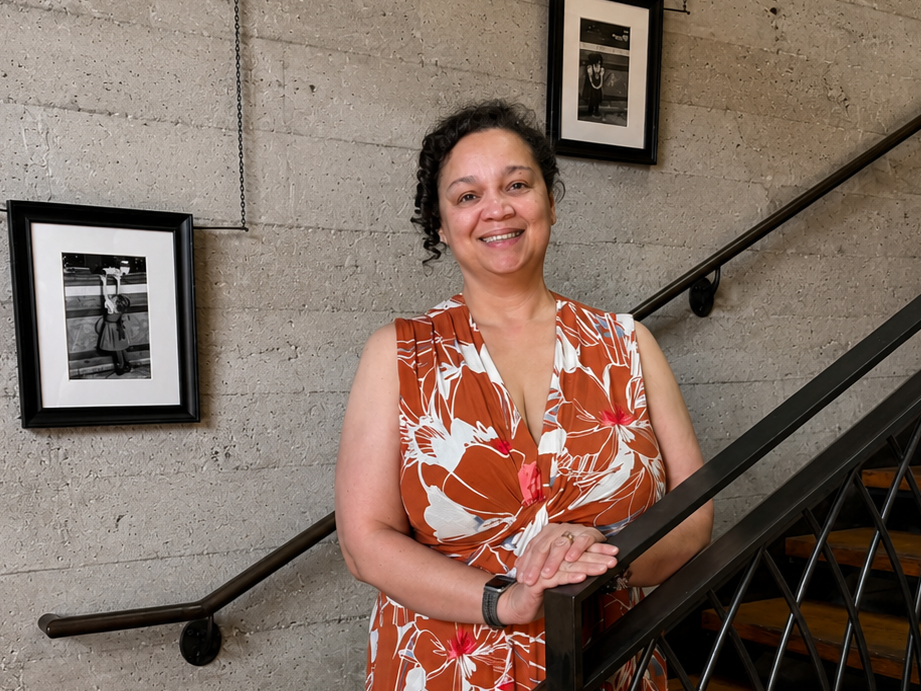 A woman smiling and standing on a staircase with a concrete wall and black and white photographs hanging on it.