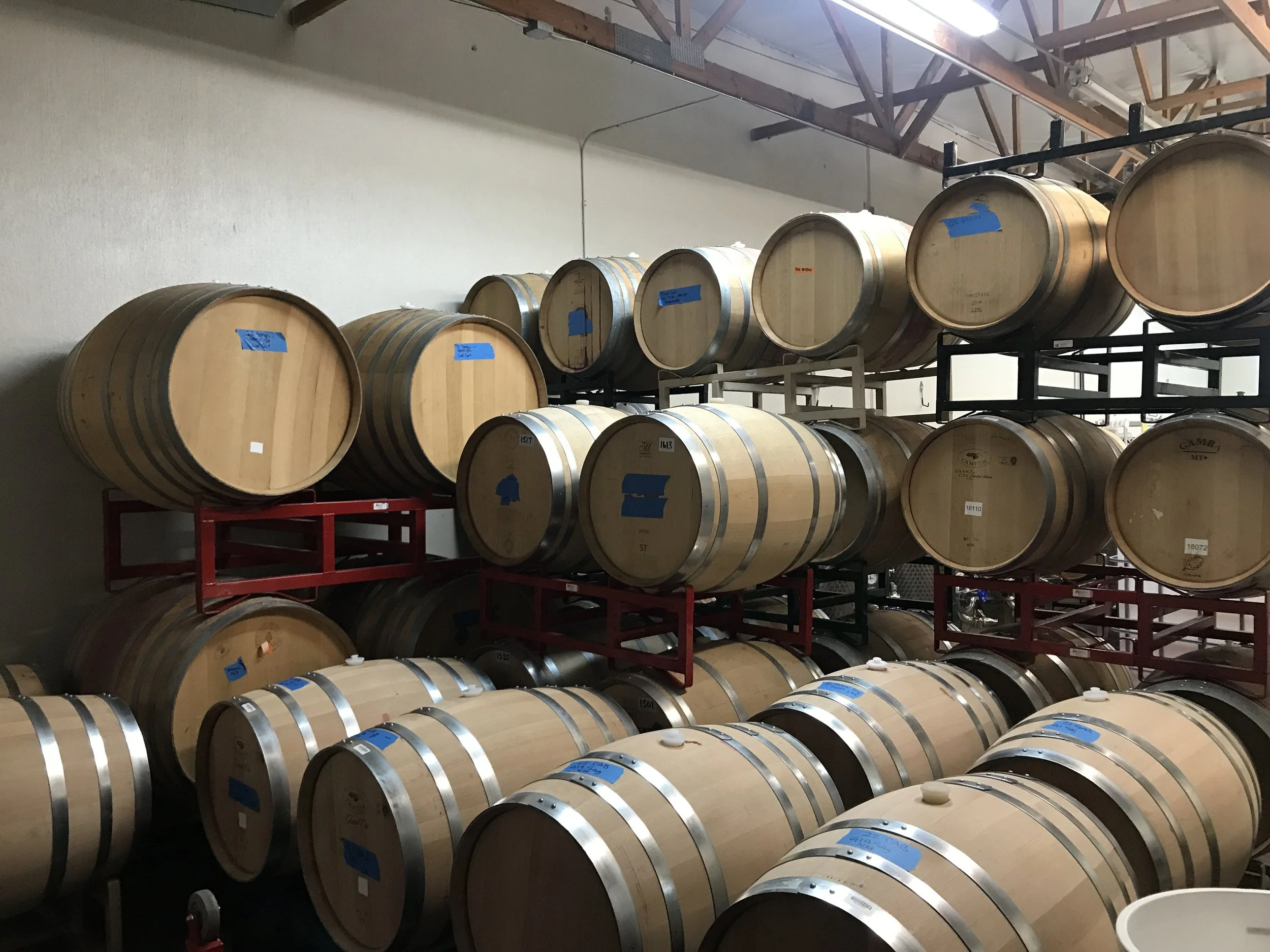 Storage room with multiple wooden barrels on metal racks, some with blue labels, in a brewery or winery.