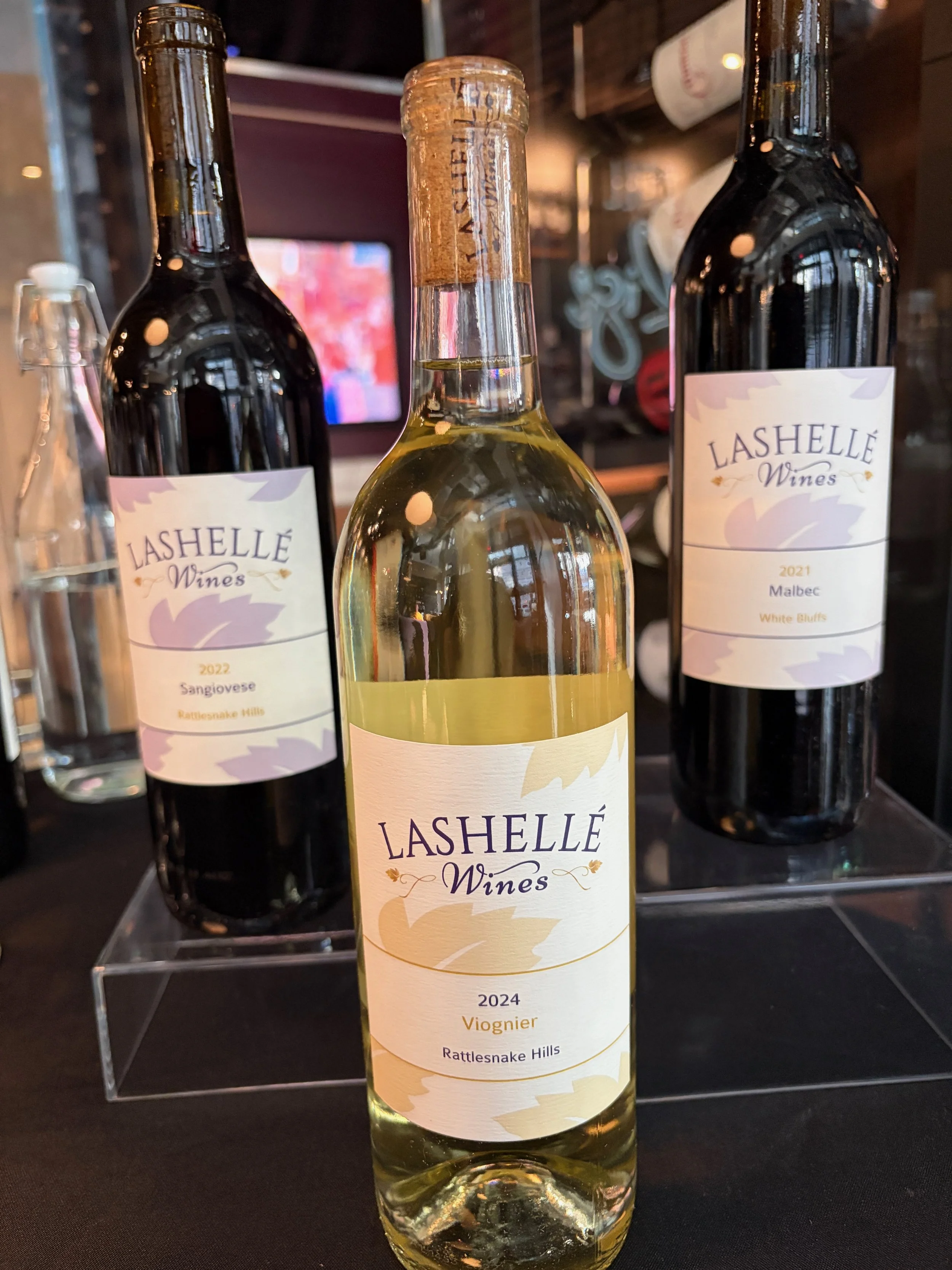 A bottle of white wine labeled 'Lashellé Wines 2024 Viognier' with two additional wine bottles in the background, placed on a dark surface.