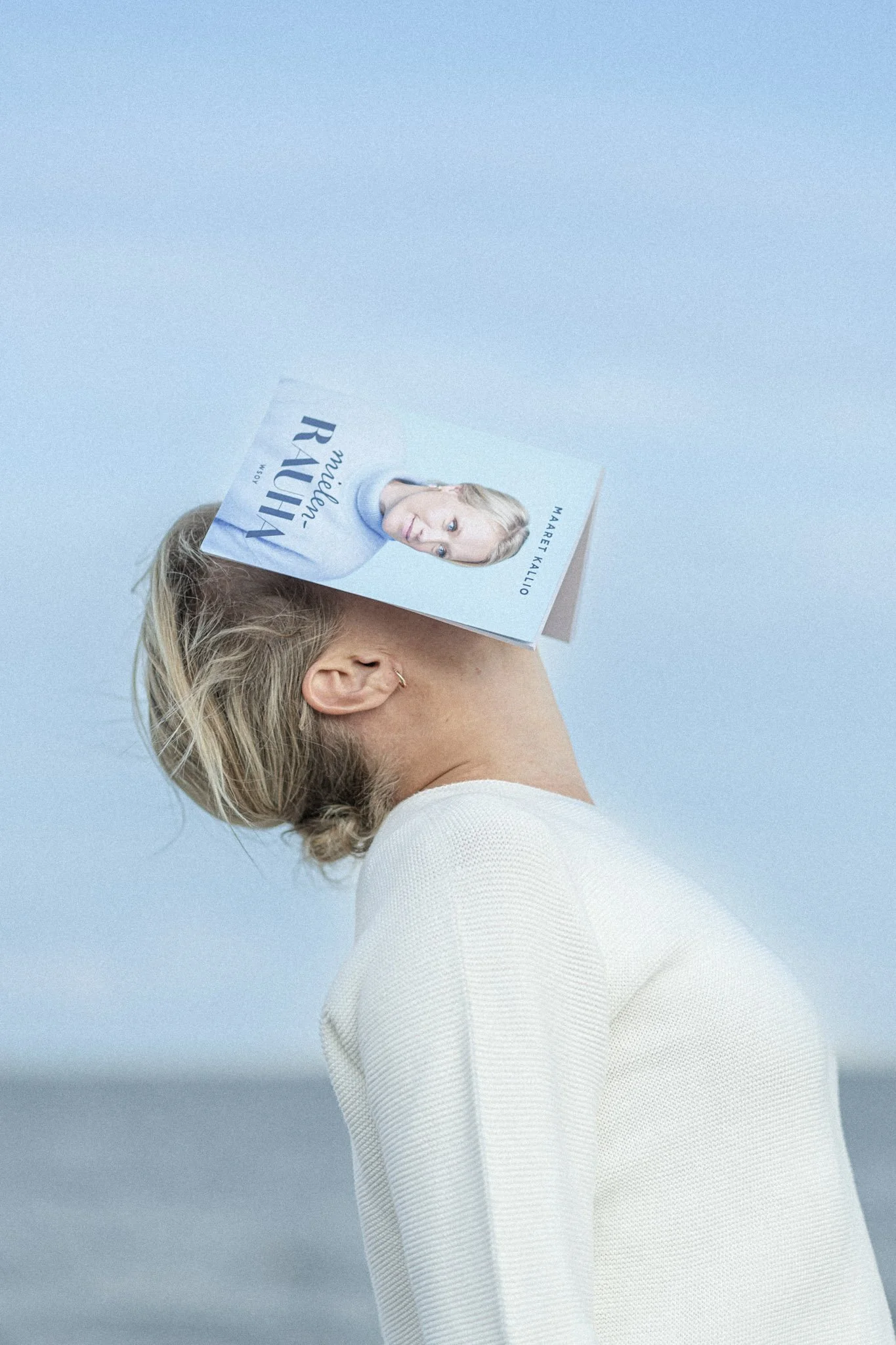 Kuvassa nainen, jolla on puolipitkä vaaleatukka, pukeutunut valkoiseen neuleeseen, ja hänellä on kirja kasvoillaan, katse suoraan kameraan, taustalla sininen taivas.