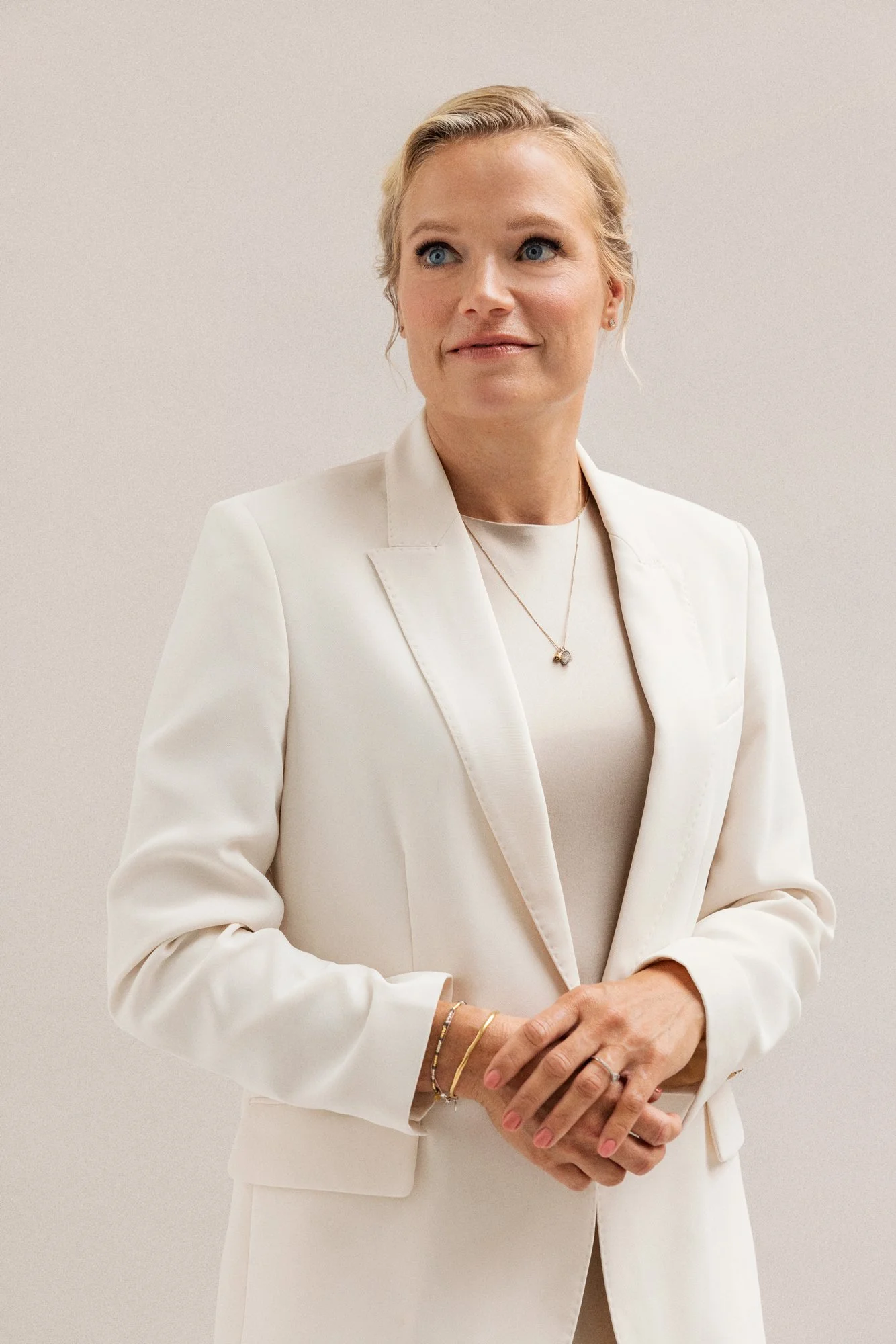 Nainen puvun tai bleiserin kanssa, vaaleat hiukset, tausta vaalea seinä.