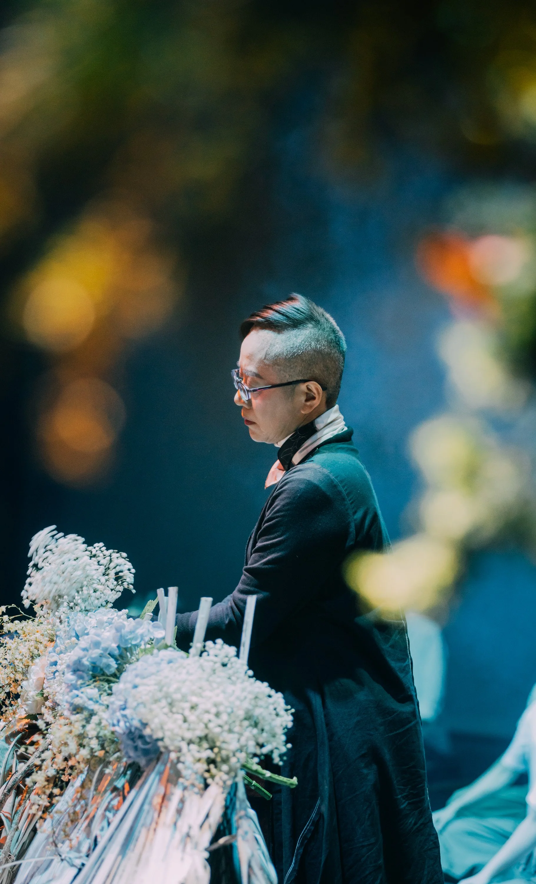 Person with glasses and a mohawk hairstyle, wearing a dark jacket, standing near a floral arrangement during a nighttime event.