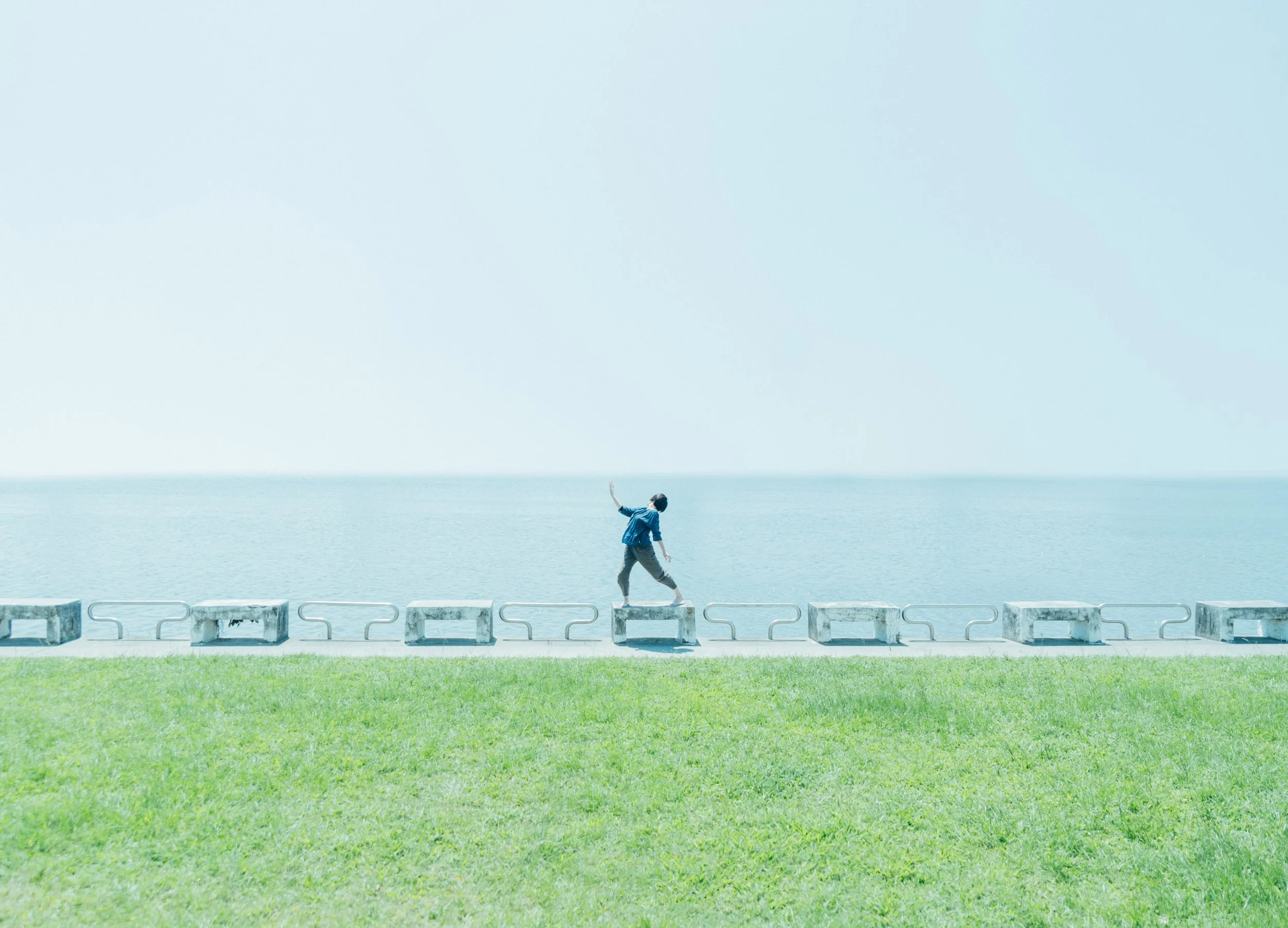 A boy in a blue jacket walking along concrete blocks by a body of water, with a clear sky overhead.