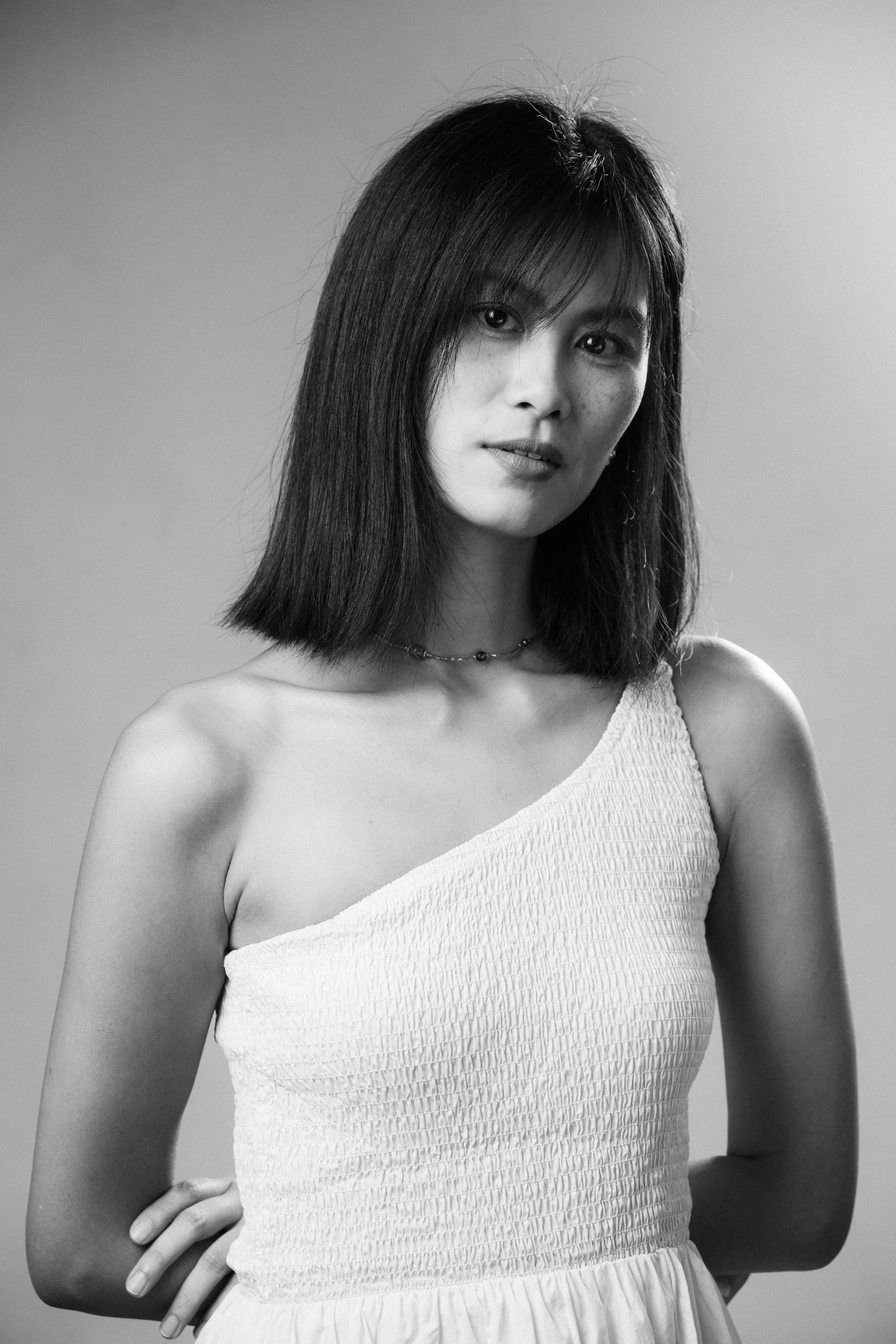 Black and white portrait of a woman with straight medium-length hair wearing a one-shoulder textured dress and a necklace, standing against a plain background.
