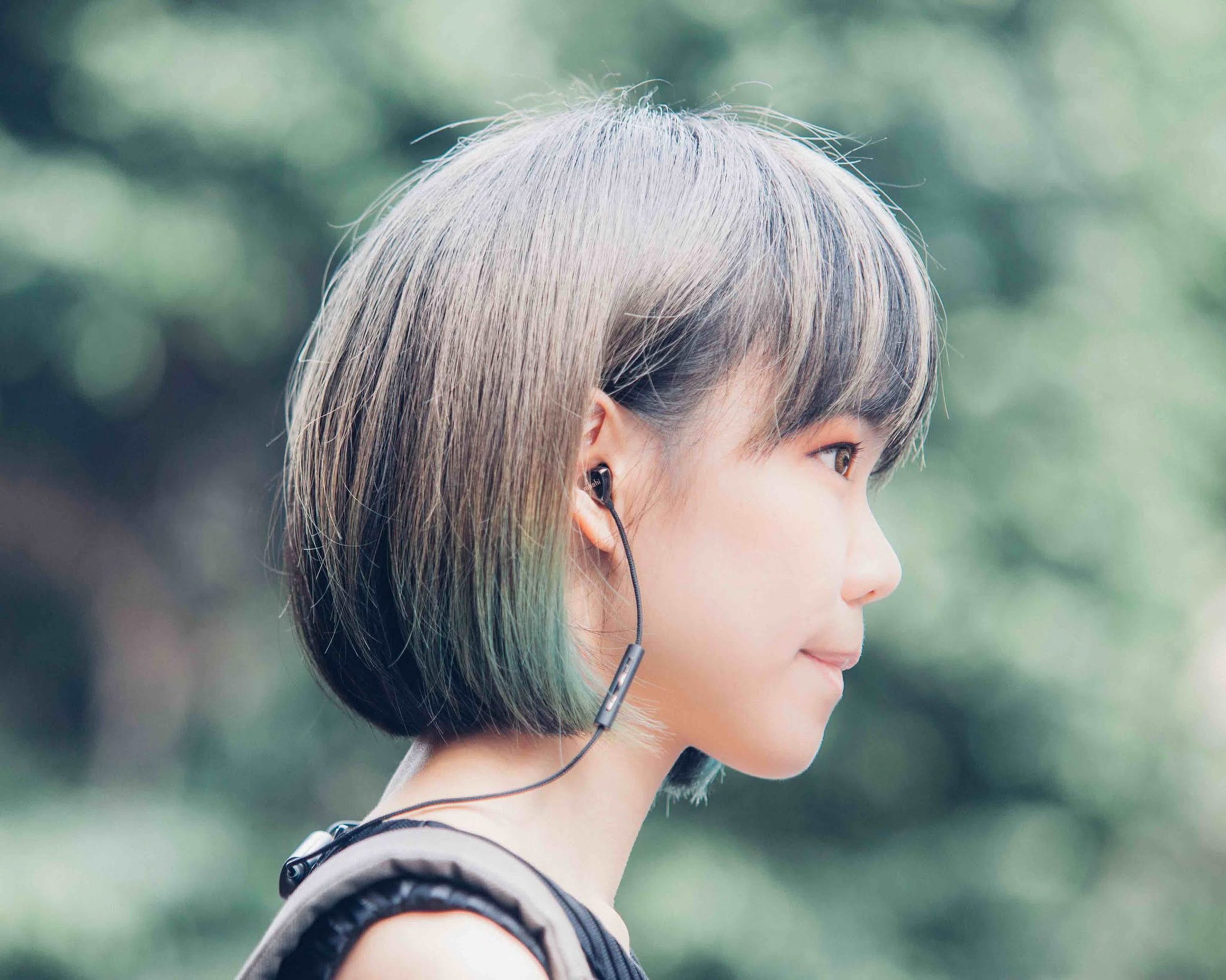 A woman with short, blonde hair wearing earbuds and a black headband, captured in profile outdoors with a blurred green background.