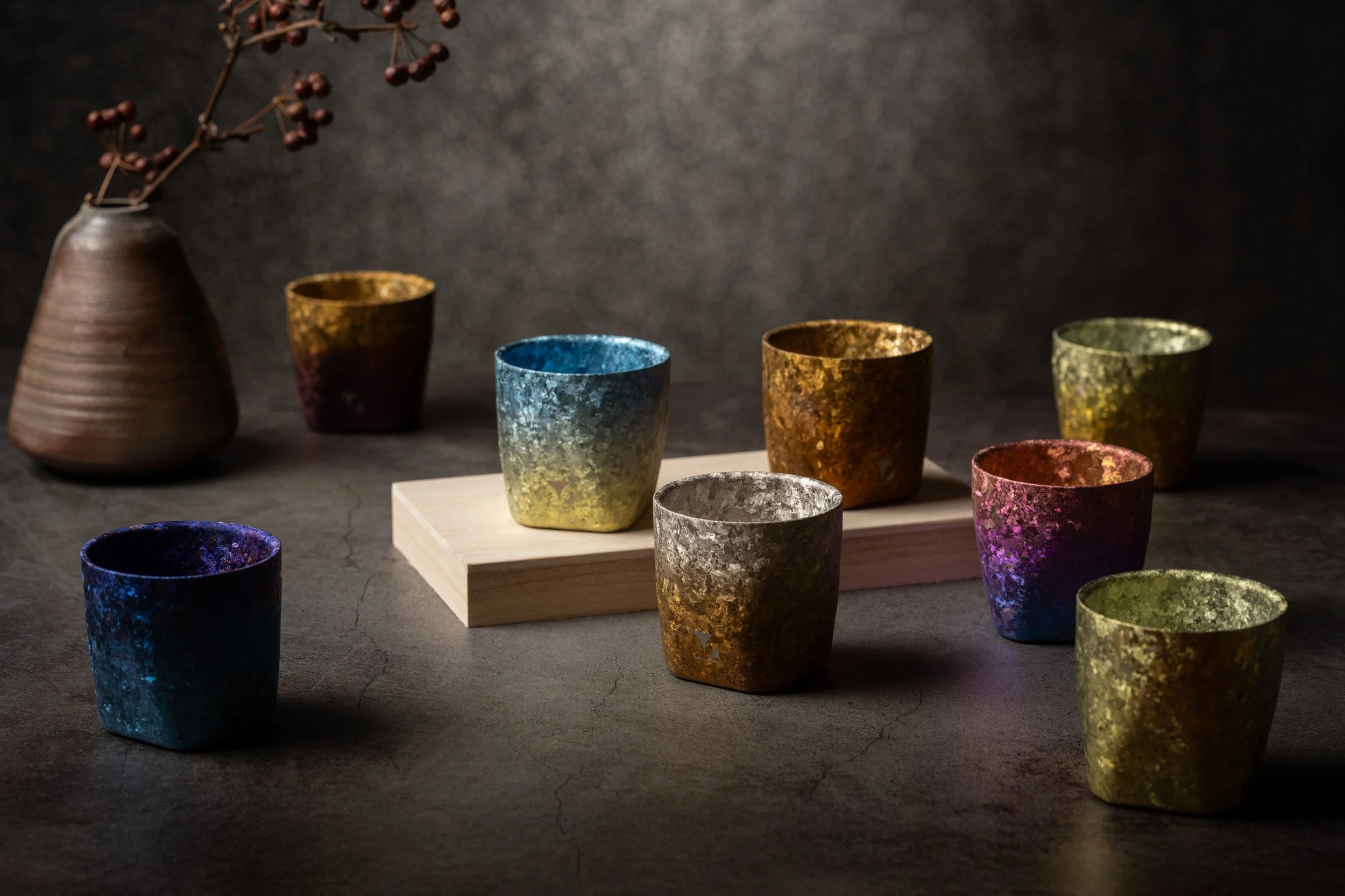 Colorful textured cups arranged on a white platform with a brown vase holding a branch with small red berries in the background.