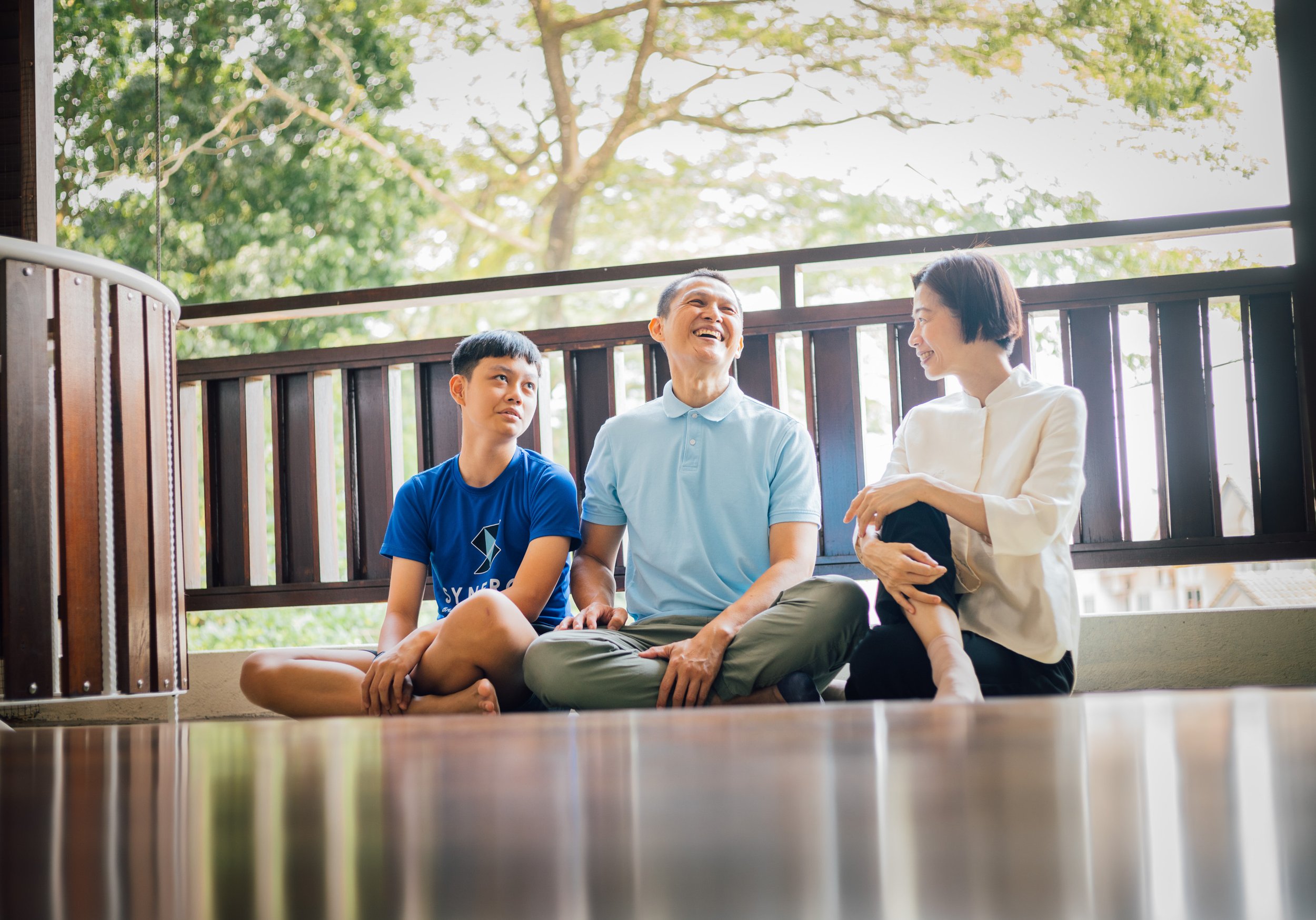An Asian family sitting on a porch, smiling and talking, with green trees and sunlight in the background.