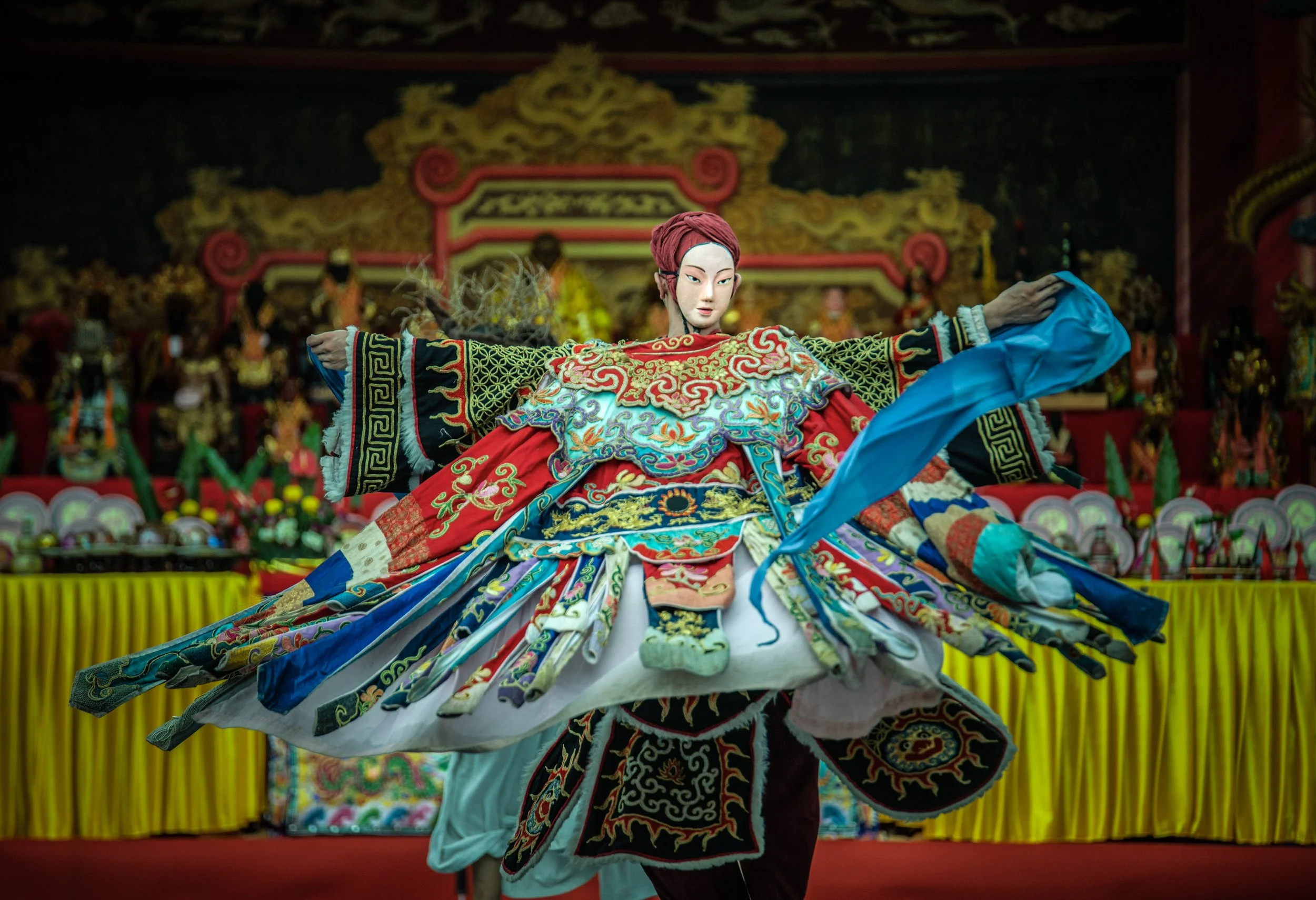 Traditional Asian puppet or performer in colorful costume with intricate embroidery, performing on stage with a dark background and decorated stage elements.