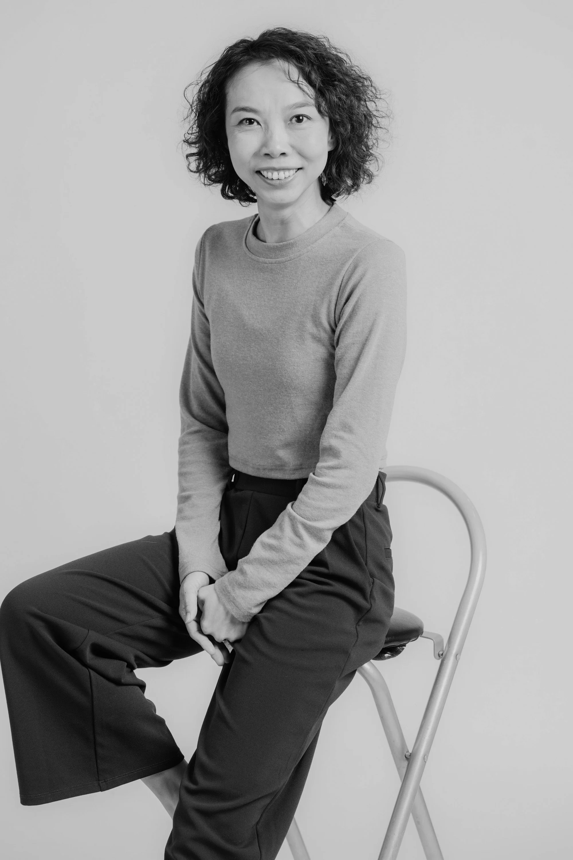 Black and white photo of a woman sitting on a chair, smiling, with curly hair, wearing a long-sleeve shirt and trousers, against a plain background.