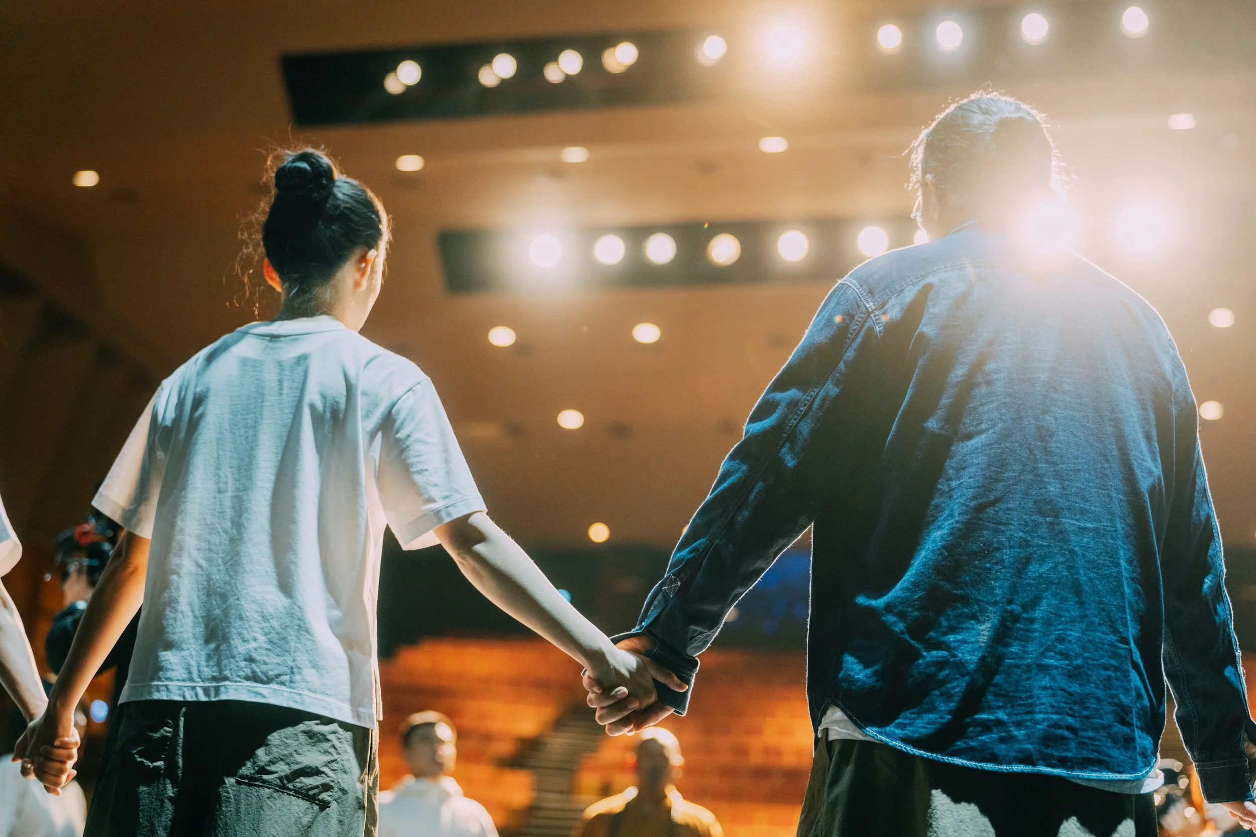 Two people, a woman and a man, holding hands and dancing in a dimly lit auditorium or theater with bright spotlights overhead.