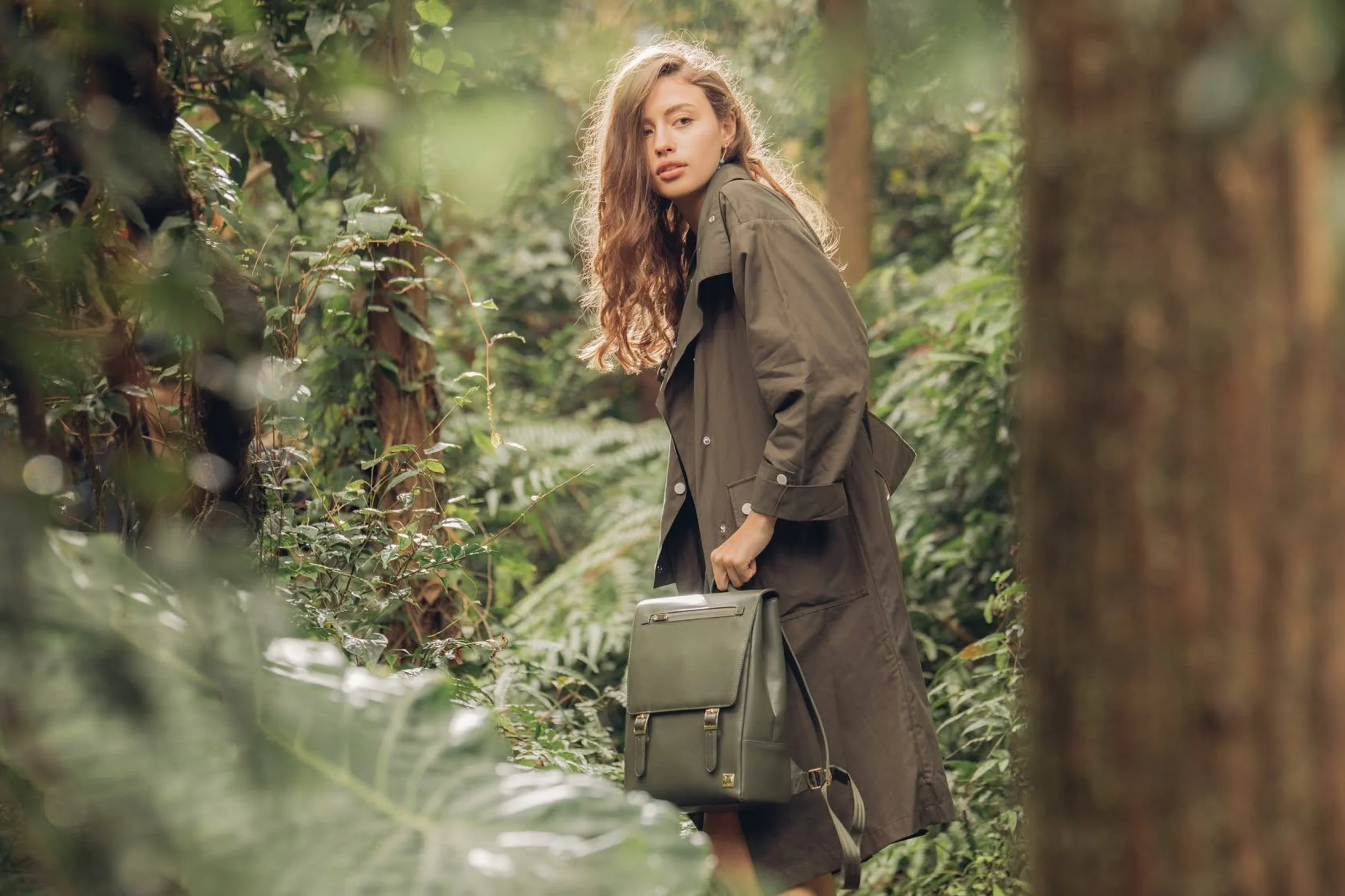 A young woman with wavy light brown hair and fair skin standing in a lush, green forest. She wears a dark green jacket and holds a matching briefcase in her right hand, looking directly at the camera with a neutral expression.