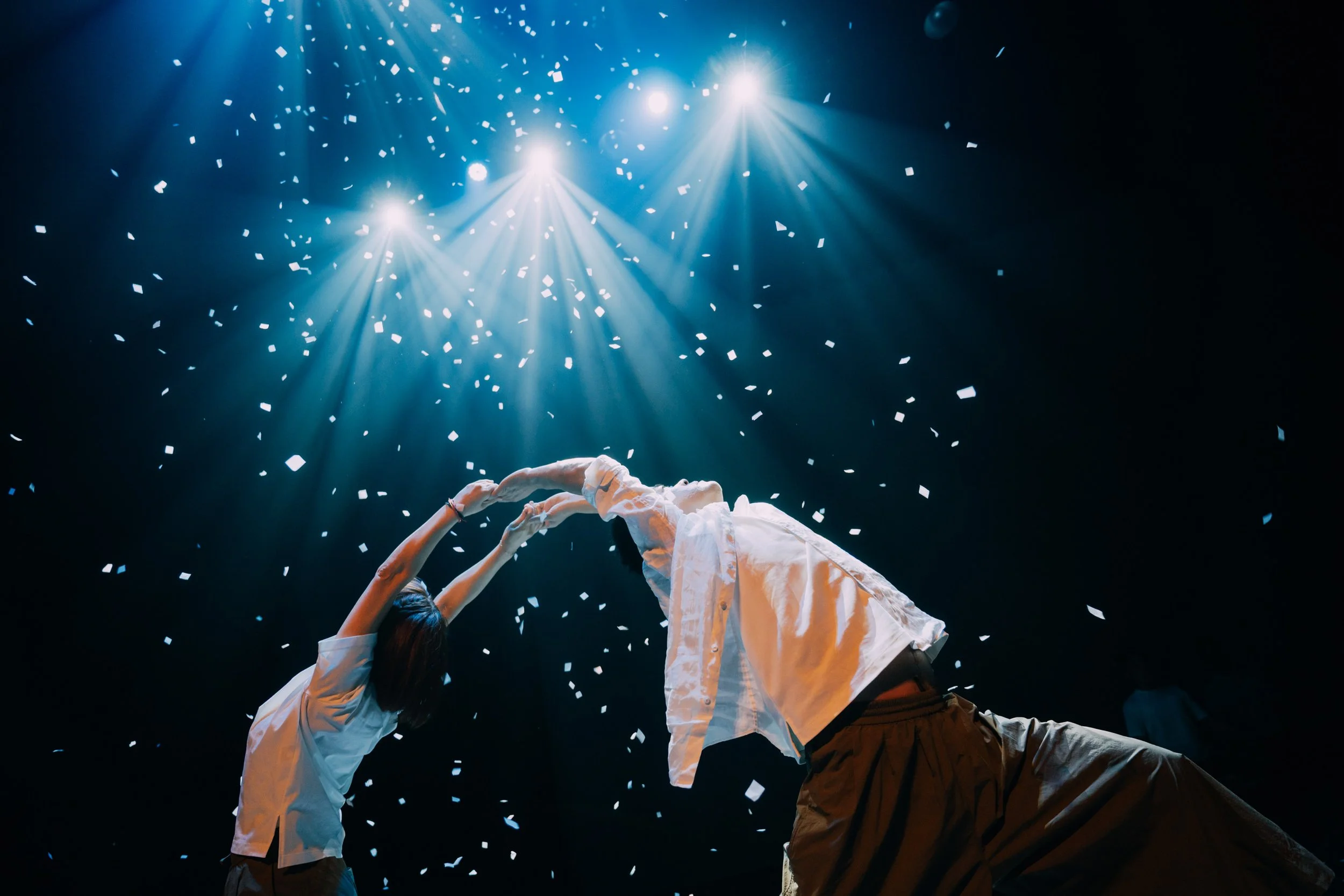 Two performers dressed in white shirts and brown pants are on stage, reaching up towards bright lights and surrounded by confetti falling from above.