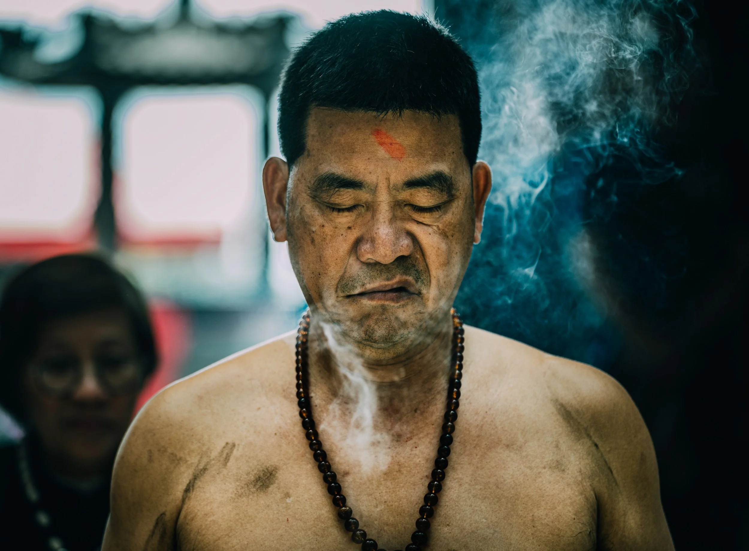 A shirtless man with closed eyes and a beaded necklace, with a red mark on his forehead and smoke surrounding him.