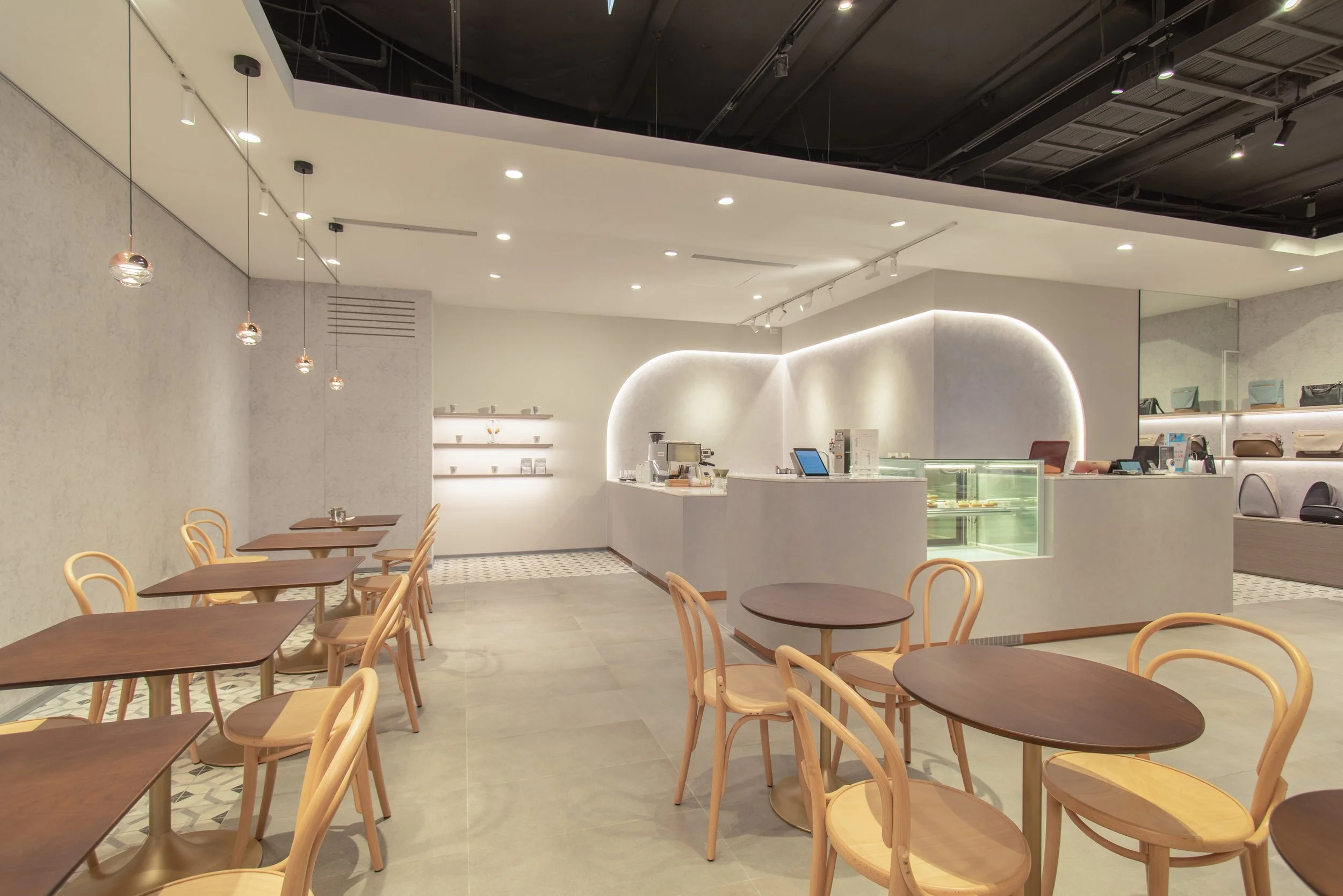 Empty modern cafe with wooden chairs around brown tables, white curved counter with display case and coffee equipment, white walls with shelves, pendant lights, and track lighting, with a dark open ceiling.