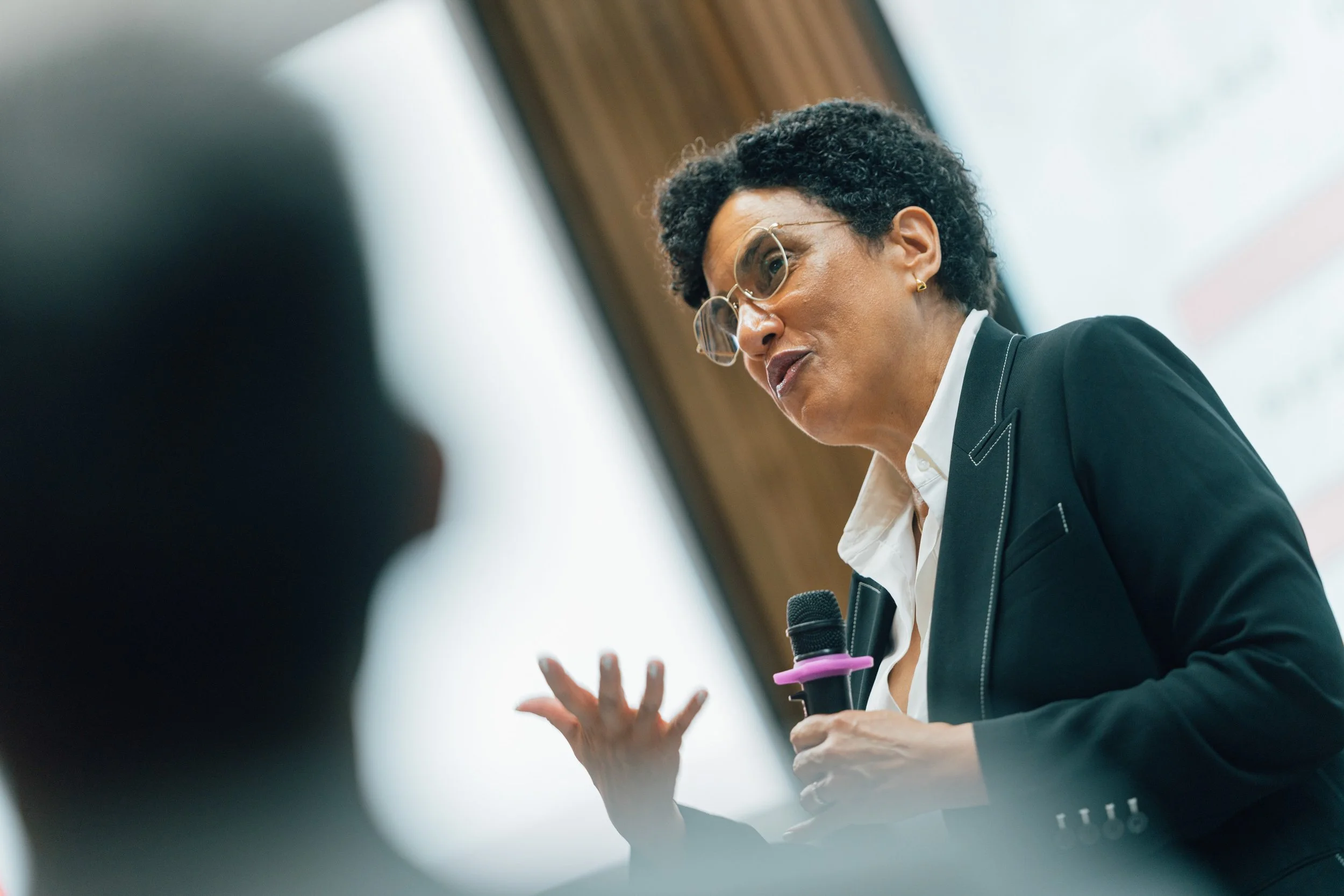 A woman with curly black hair, glasses, and earrings is speaking passionately, holding a microphone, with her hands gesturing, against a blurred background with wooden paneling and large windows.