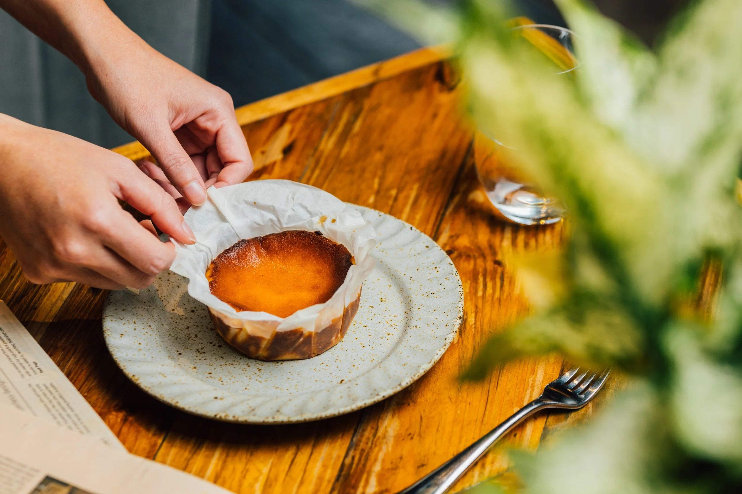 A person's hands peeling away parchment paper from a small, round, golden-brown cake or dessert on a white speckled plate, set on a wooden table with a fork and a glass of water nearby.