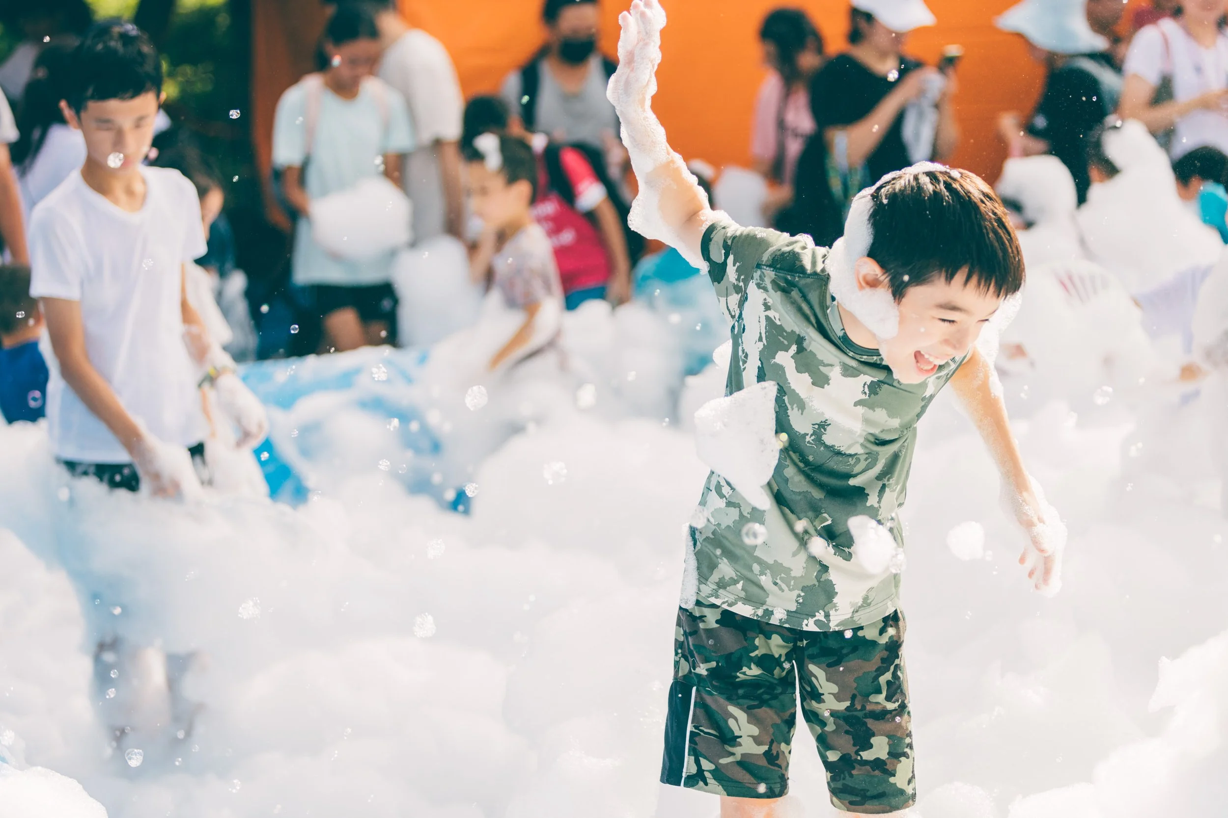 Children playing with foam or bubbles outdoors during daytime, some smiling and enjoying the activity.