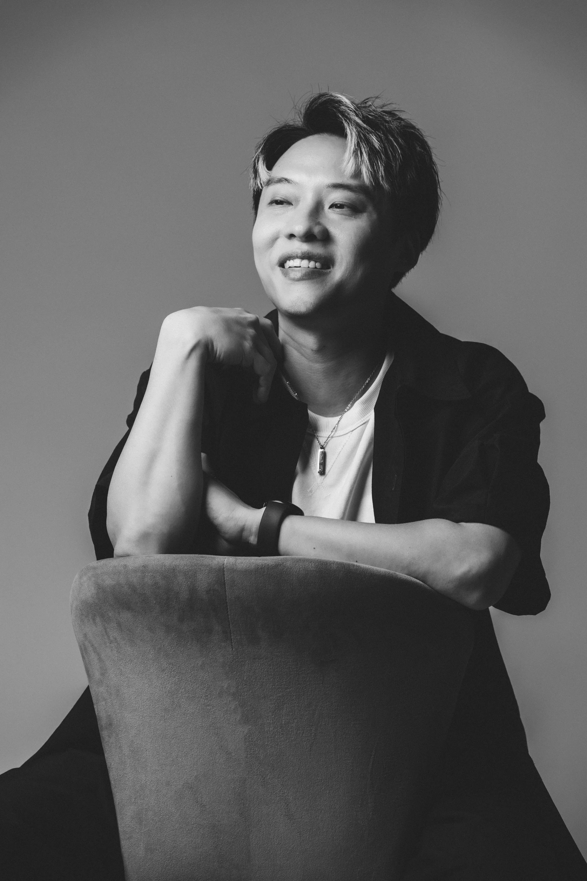 Black and white portrait of a young man with short, tousled hair, smiling with a slight gap in his teeth. He is wearing a dark jacket over a light shirt, has a necklace, a watch on his wrist, and is seated with his arm resting on a chair, with his ha