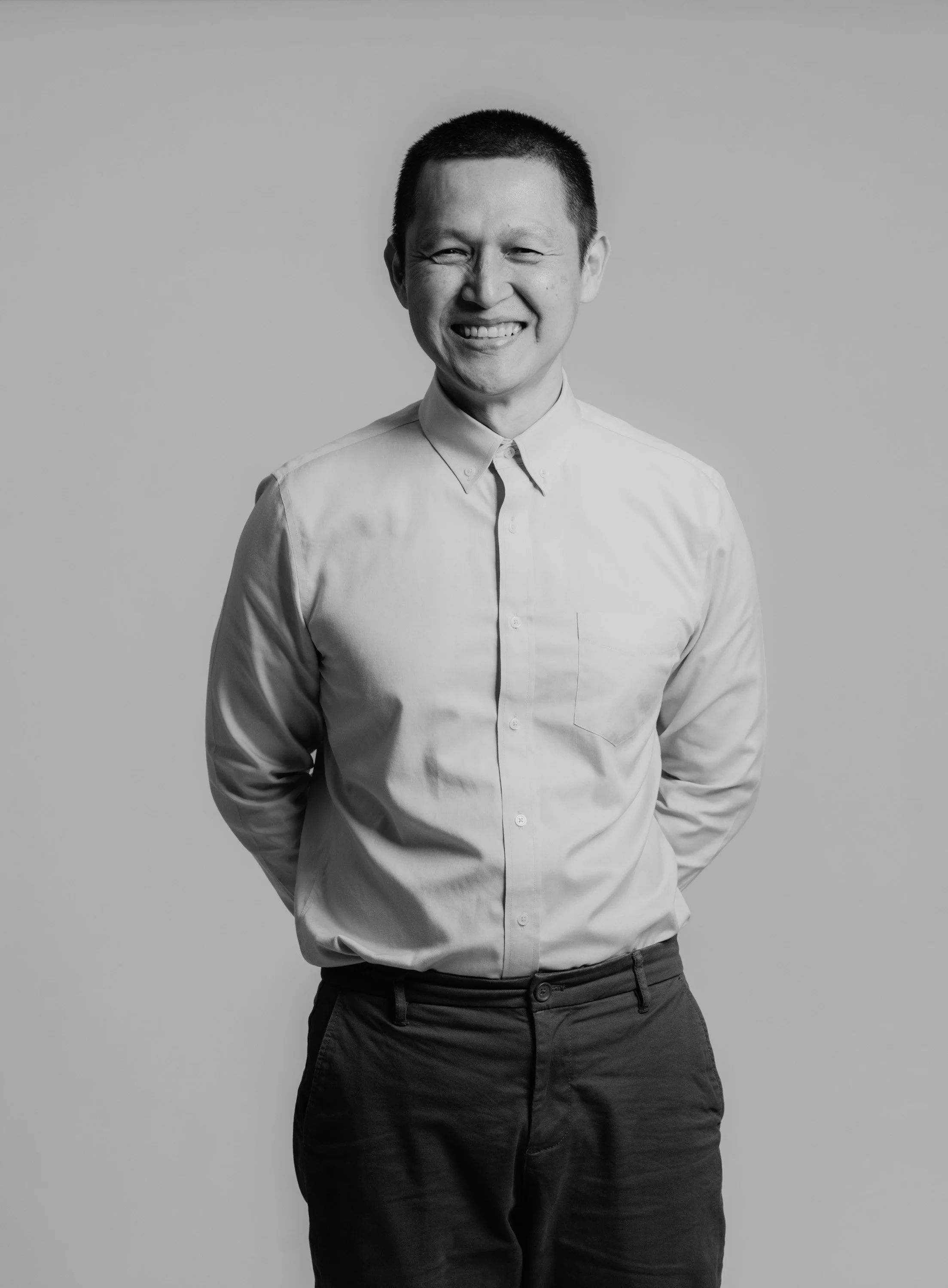 Black and white image of a smiling man with short hair wearing a button-up shirt and dark pants, standing against a plain background.