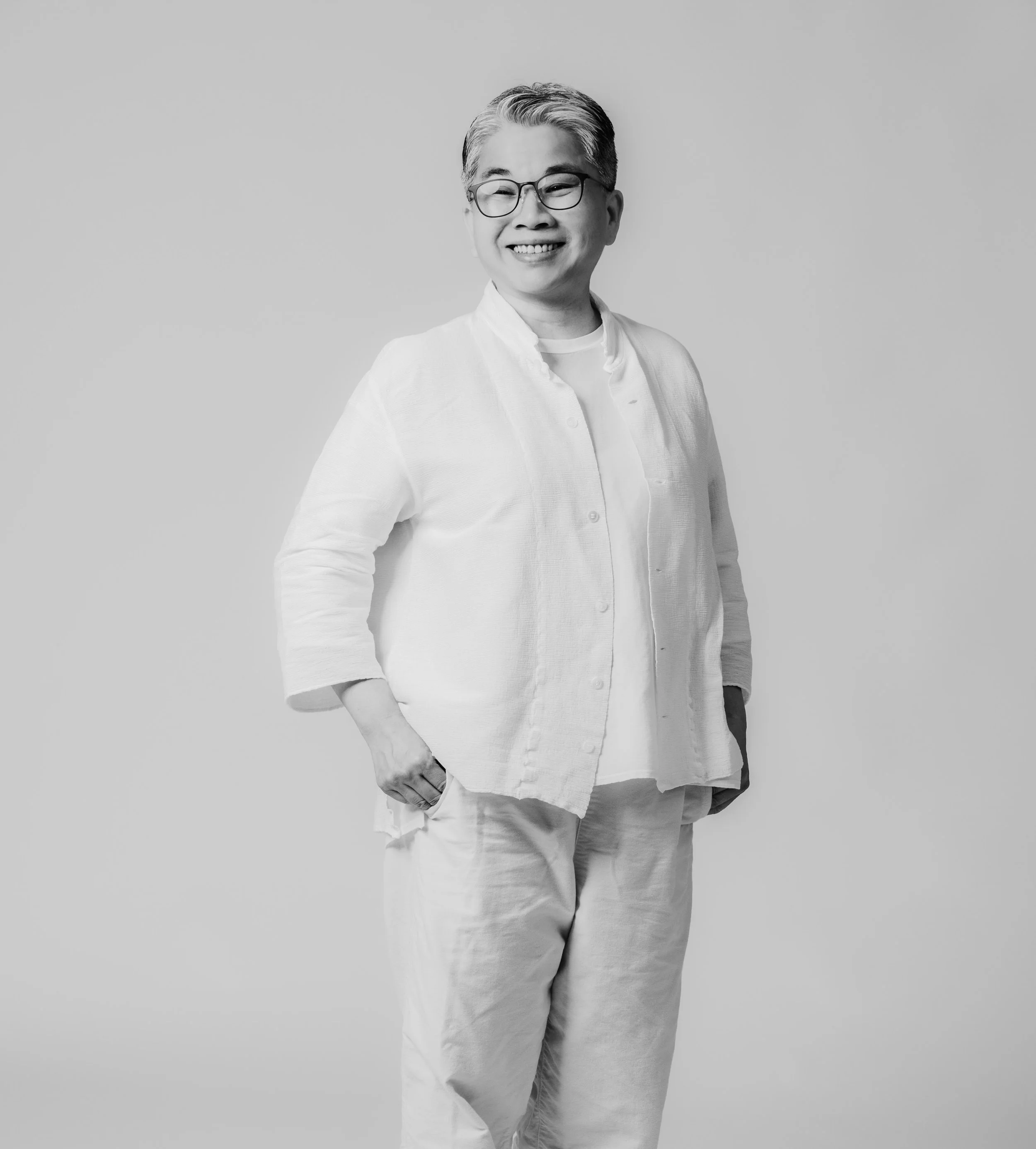 Black and white portrait of a smiling person with short, styled hair, wearing glasses, a light-colored shirt, and light-colored pants, standing against a plain background.
