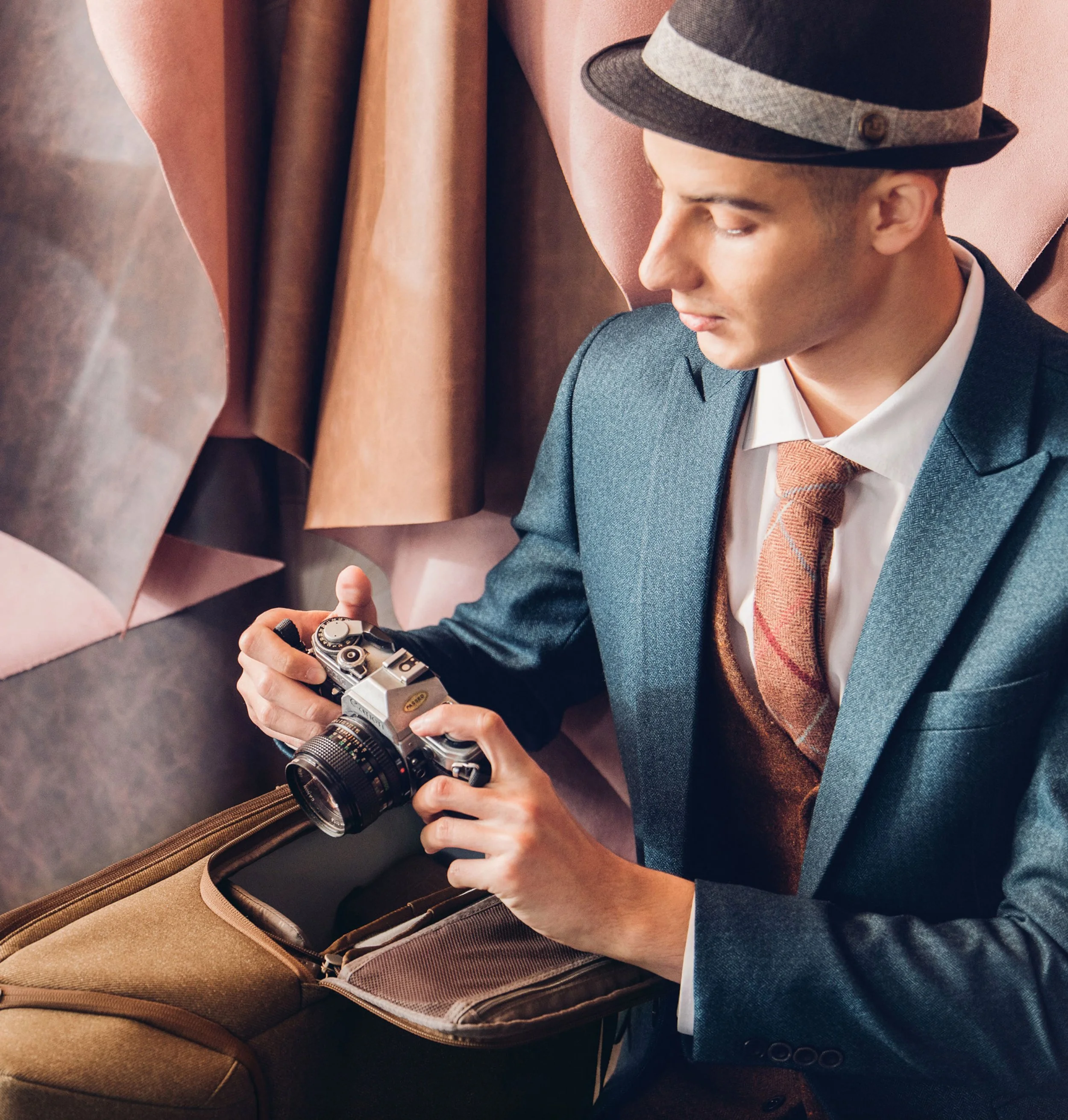 A man in a suit and tie wearing a gray fedora hat, holding a vintage film camera, standing next to a brown suitcase with emboidered brown straps and zippers, against a backdrop of hanging fabric.