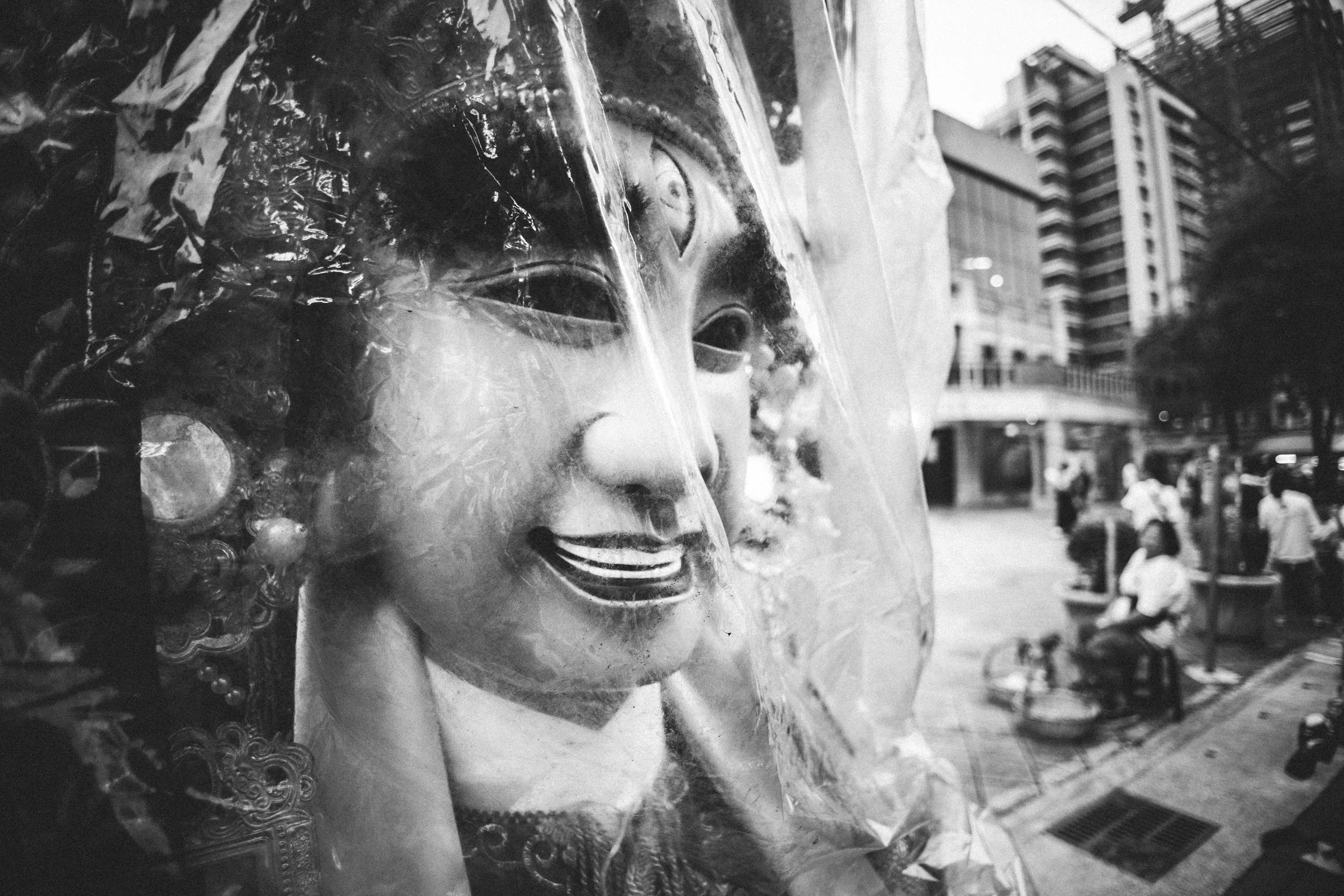 Close-up of a smiling decorative mask displayed in an urban outdoor setting with tall buildings and people in the background.