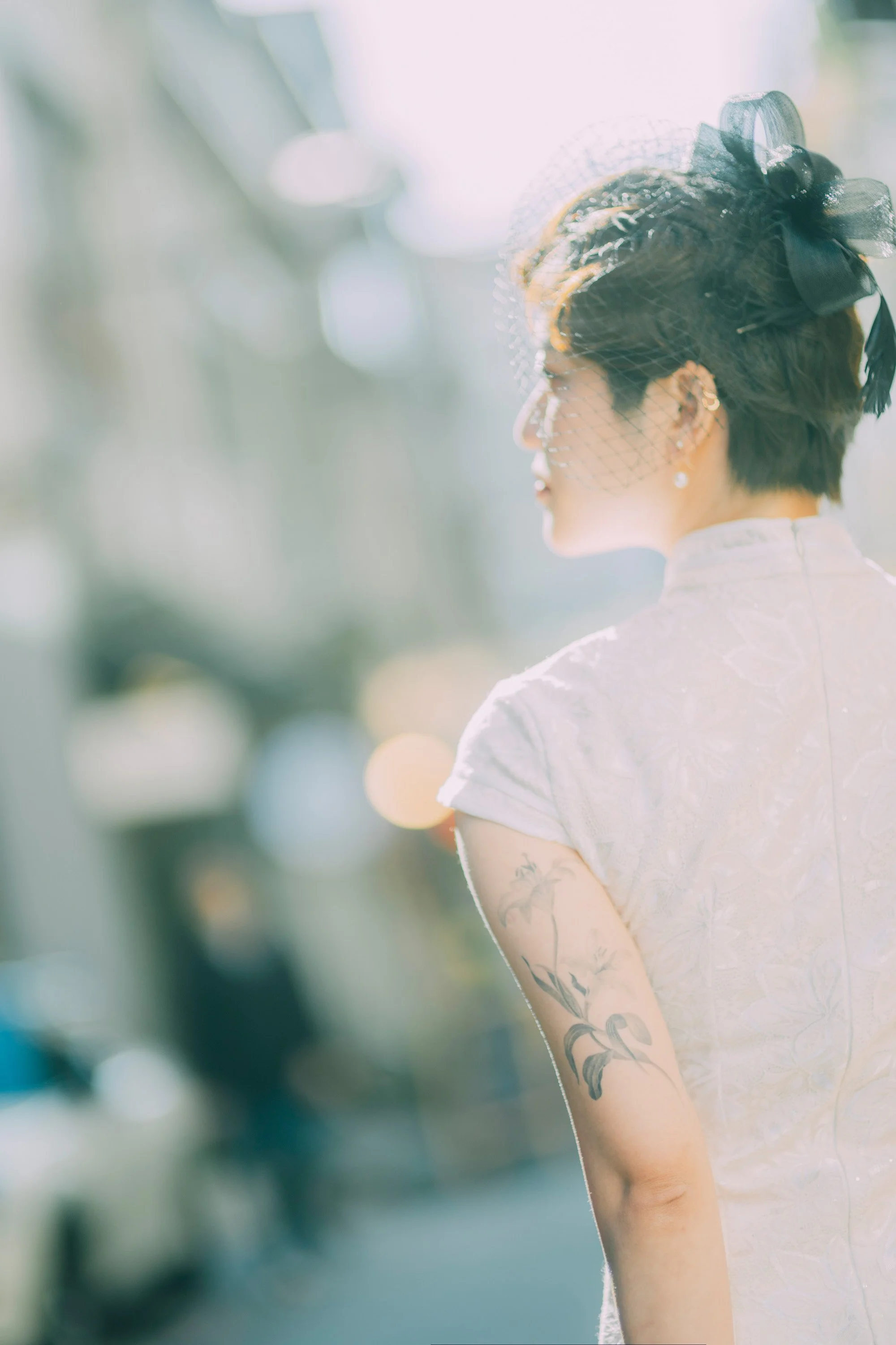 Profile of a woman with short dark hair wearing a dark lace headband and earrings, with tattoos on her arm, in bright outdoor sunlight.