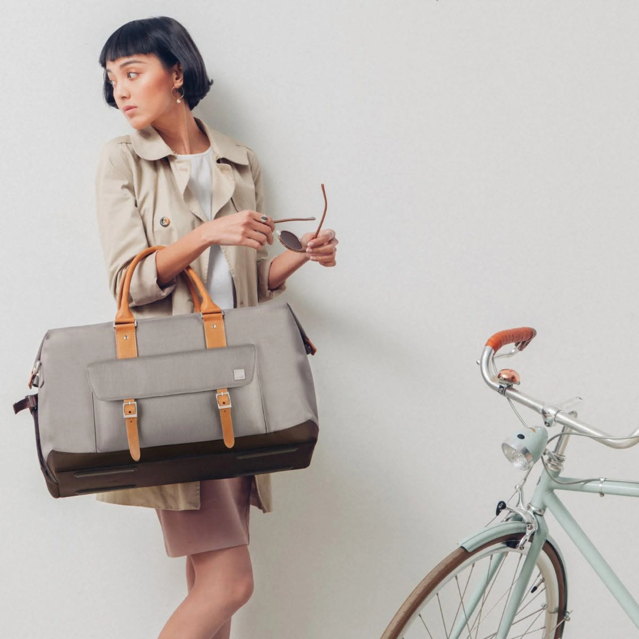 A woman with short dark hair wearing a beige trench coat holding sunglasses, standing in front of a plain white wall near a mint green bicycle.