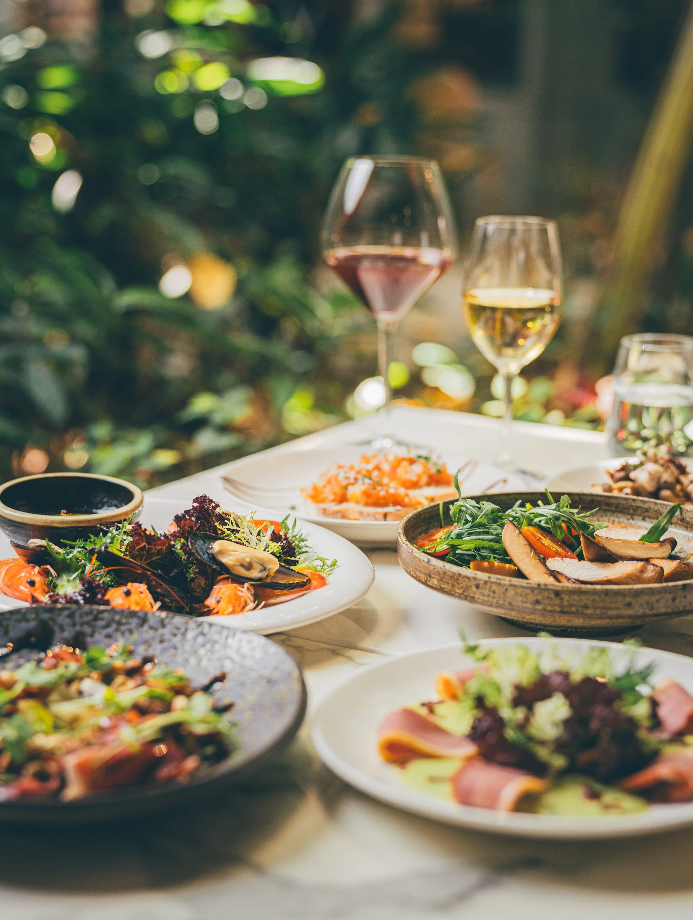 A table set with various dishes including salads and a glass of red wine, with a blurred background of greenery.