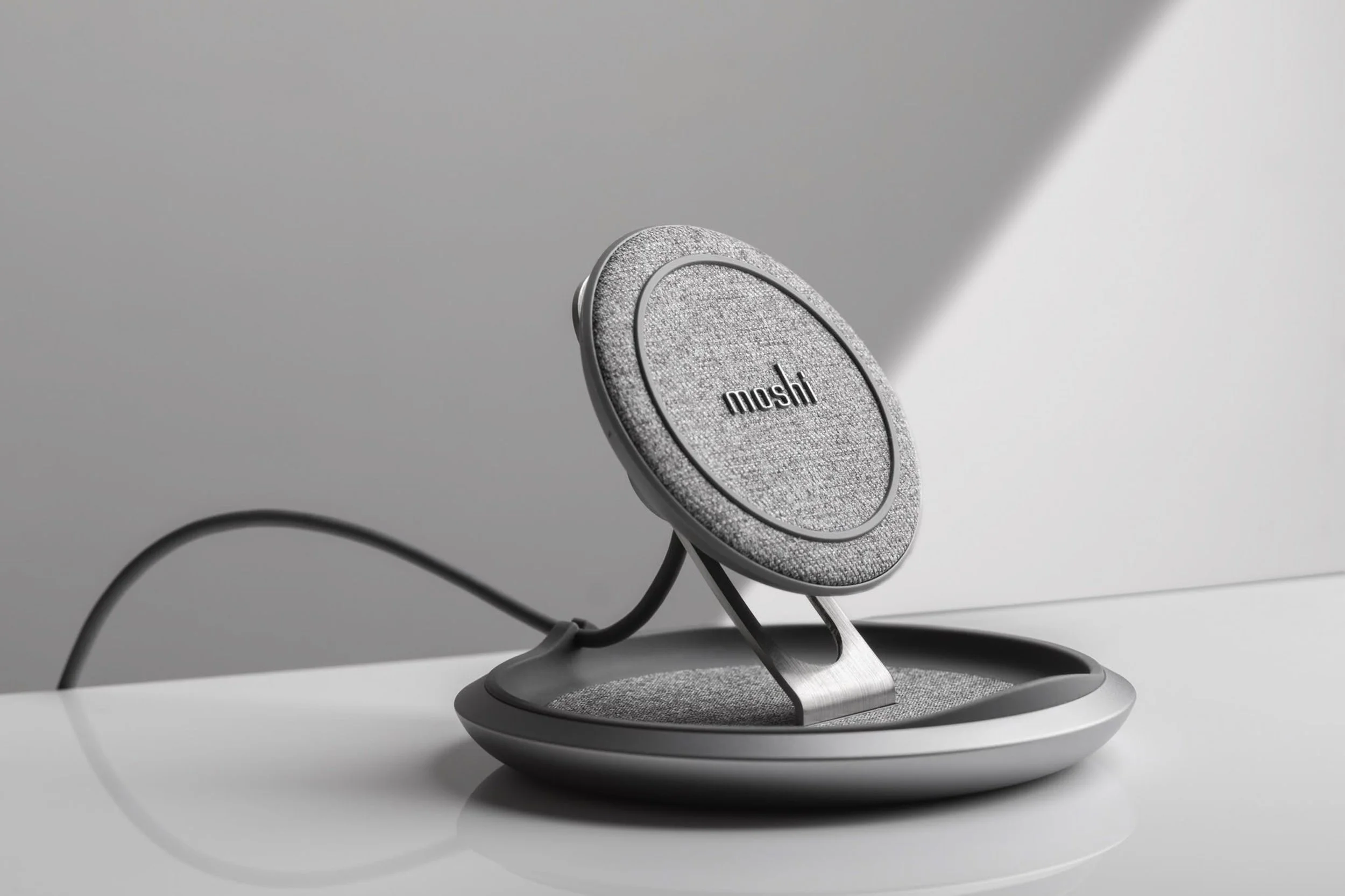 A close-up view of a silver, fabric-covered wireless charging pad with the brand name 'moshi' on it, connected by black and gray cables on a white surface.