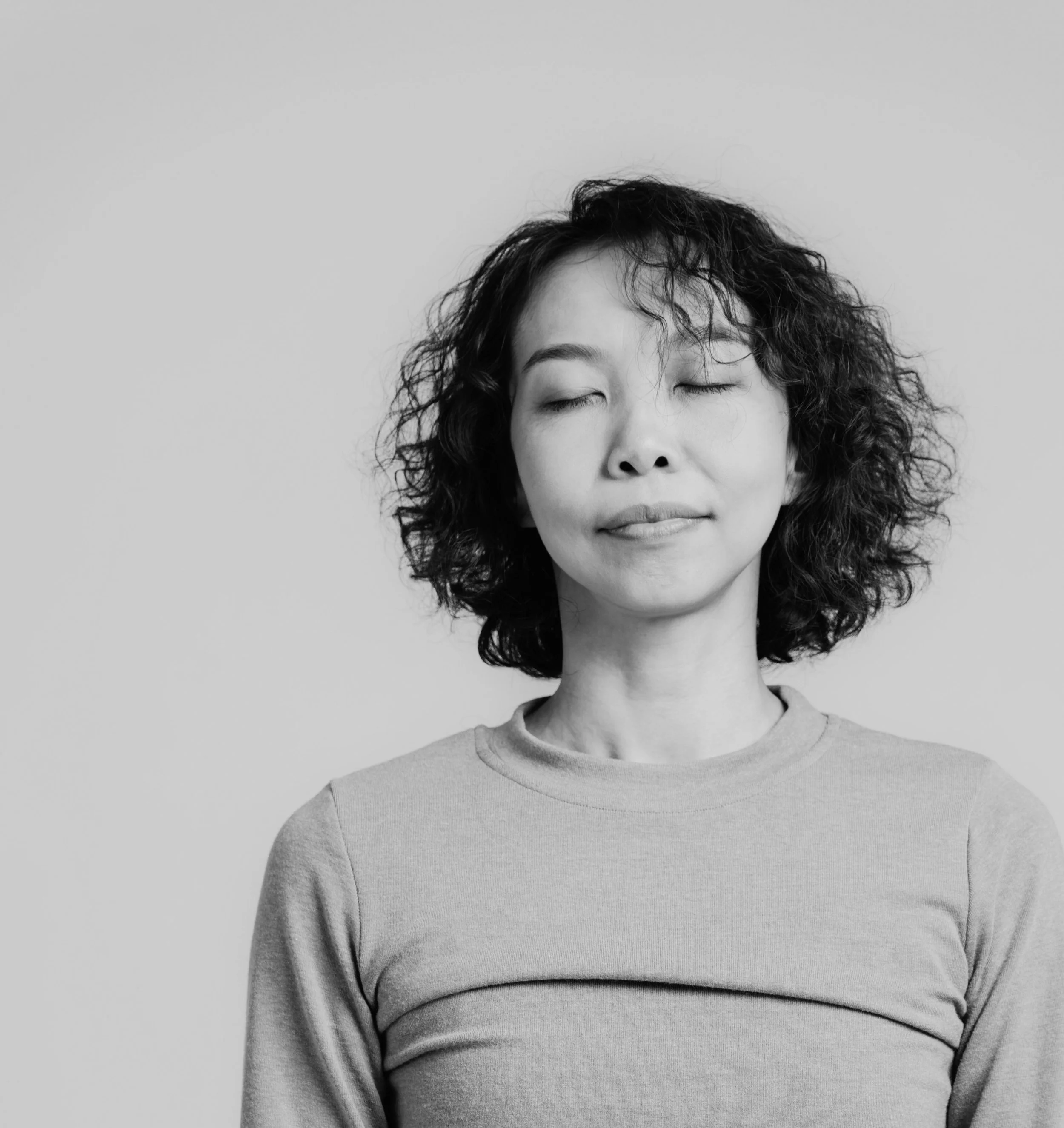 Close-up of a person with curly hair and closed eyes, wearing a light-colored top, against a plain background.