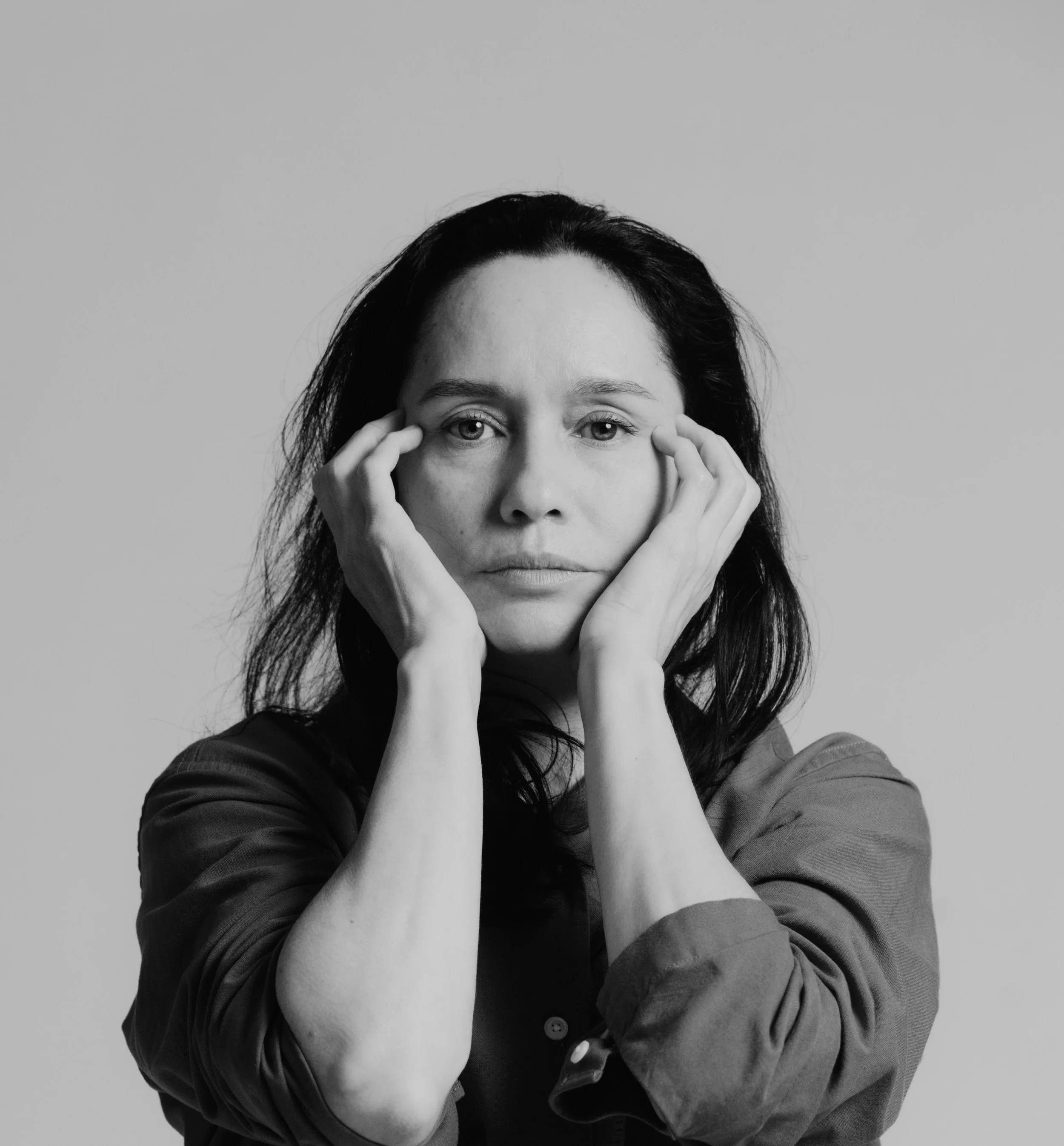A black and white portrait of a woman with dark hair, holding her face with both hands, looking directly at the camera against a plain background.