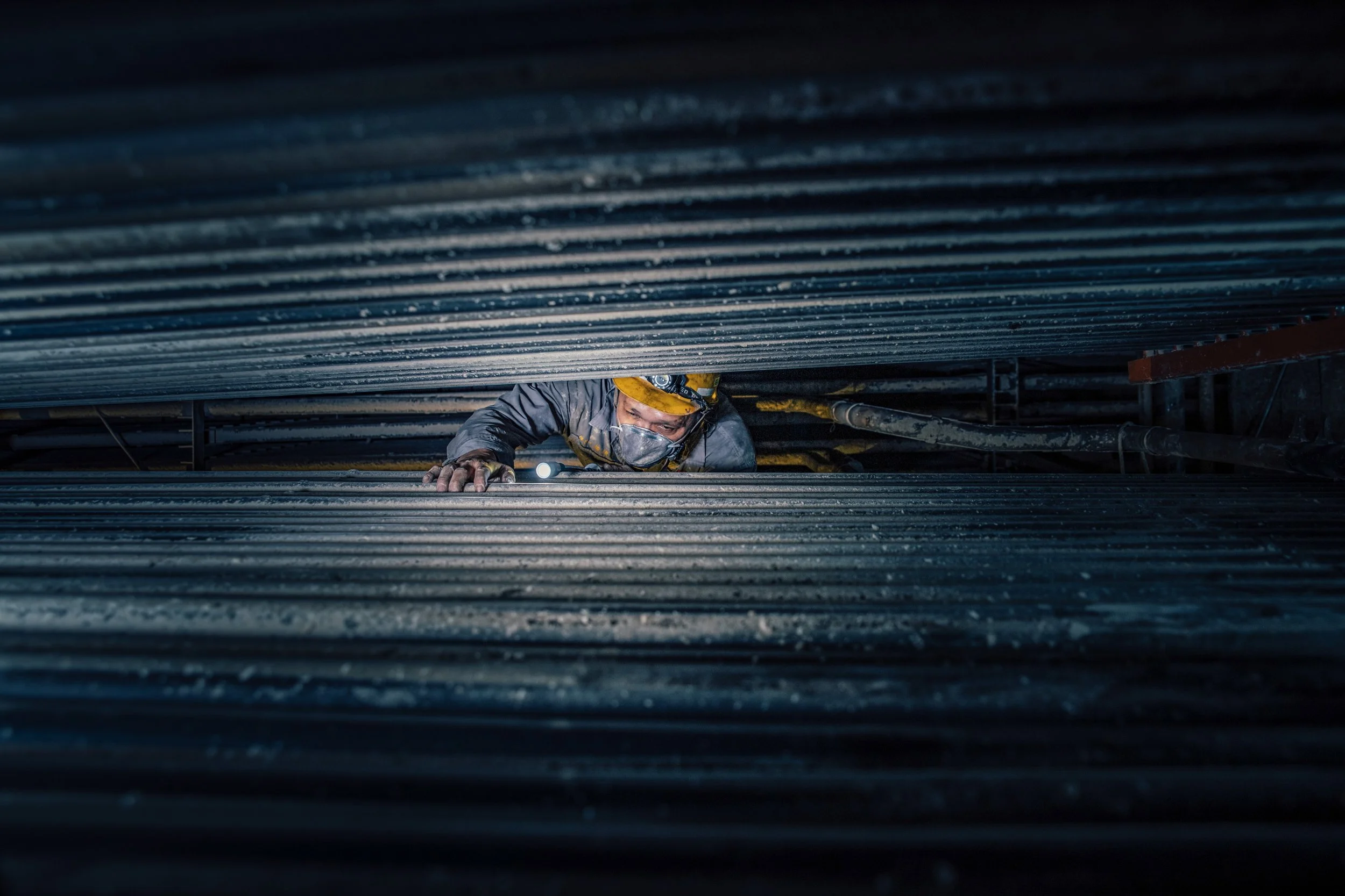 Close-up view of a worker in a hard hat and face mask crawling on a ledge under a metal structure, with dark surroundings.