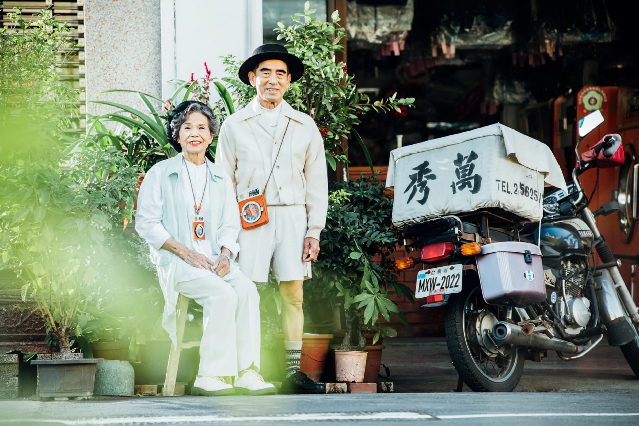 An elderly Asian couple standing outdoors near potted plants and a parked motorcycle with a covered cargo box. The woman is seated and wearing white, the man is standing and wearing a light-colored jacket and hat, both smiling.
