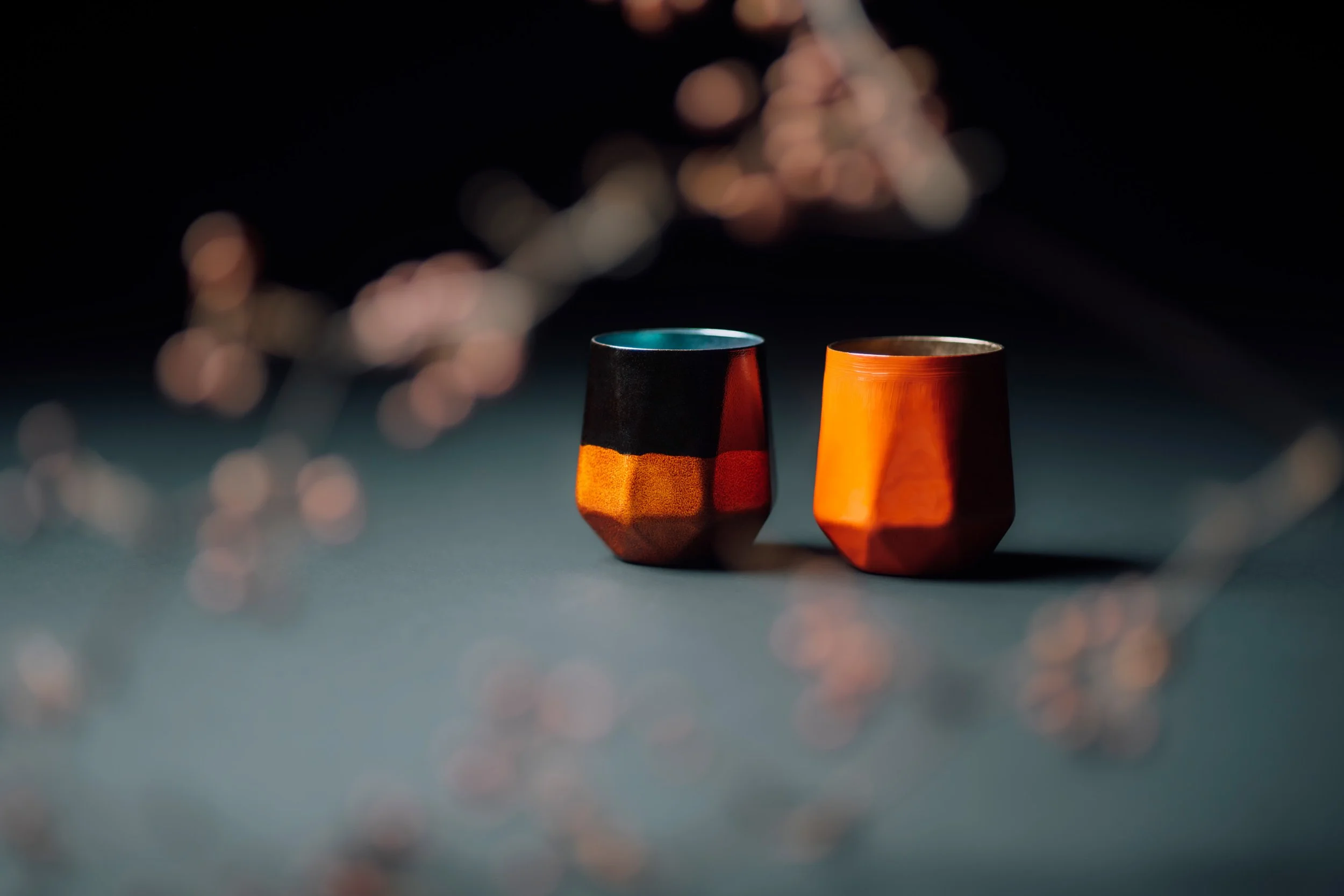 Two colorful cups, one black and orange, one orange, sitting on a surface with blurred bokeh lights in the background.