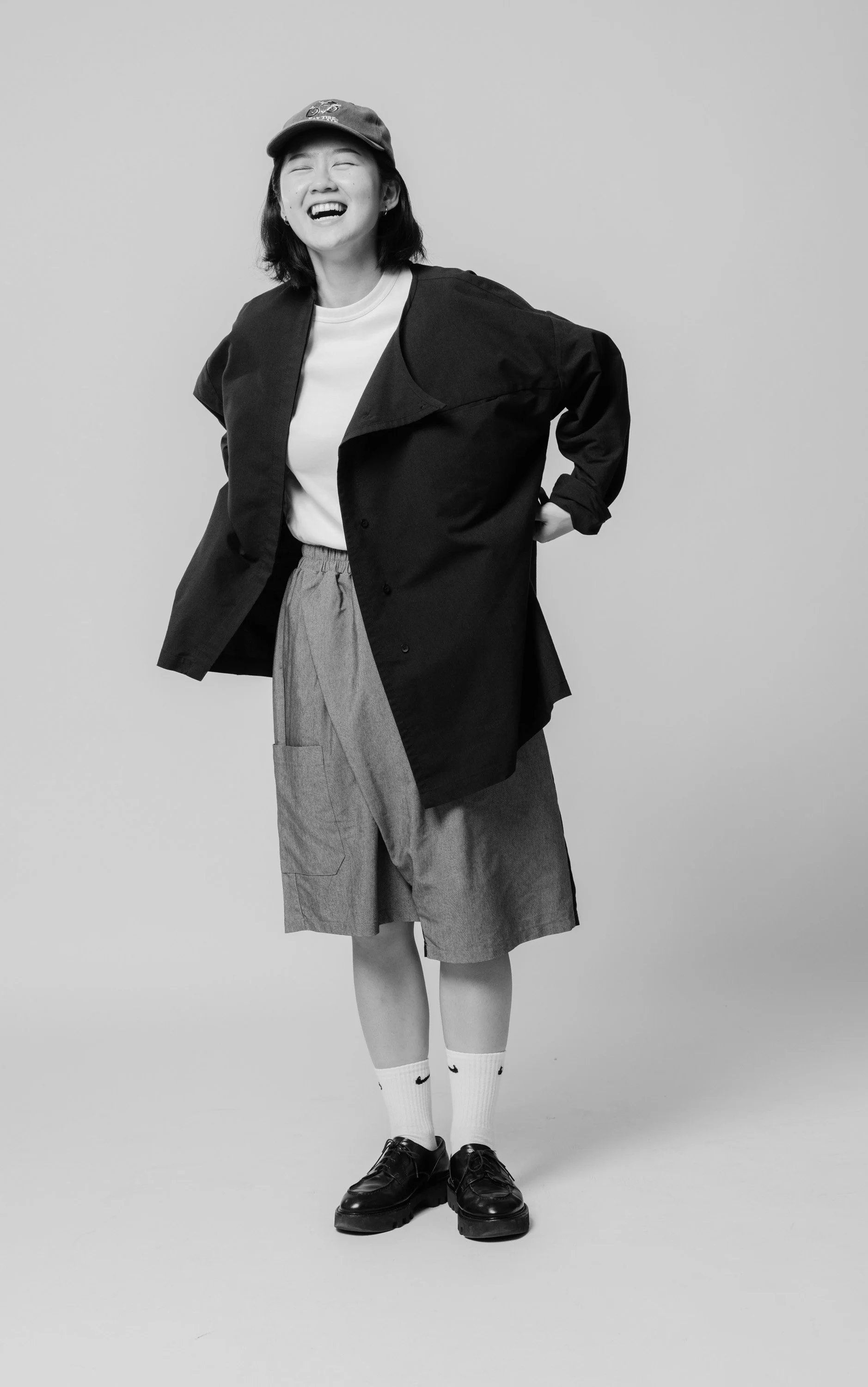 Black and white photo of a young woman laughing, wearing a baseball cap, white T-shirt, oversized dark jacket, knee-length shorts, white socks, and black shoes, standing against a plain light gray background.